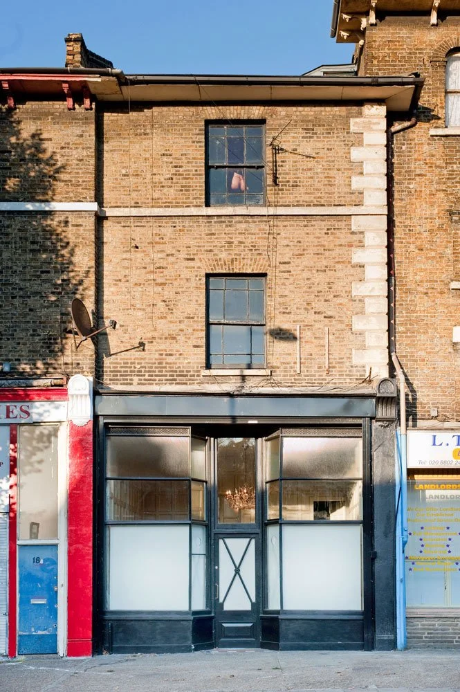 A three-story brick building with a storefront on the ground level. The store has large frosted glass windows and a black entrance door. The upper two floors have two windows each, and the building shows some signs of age with a satellite dish attach