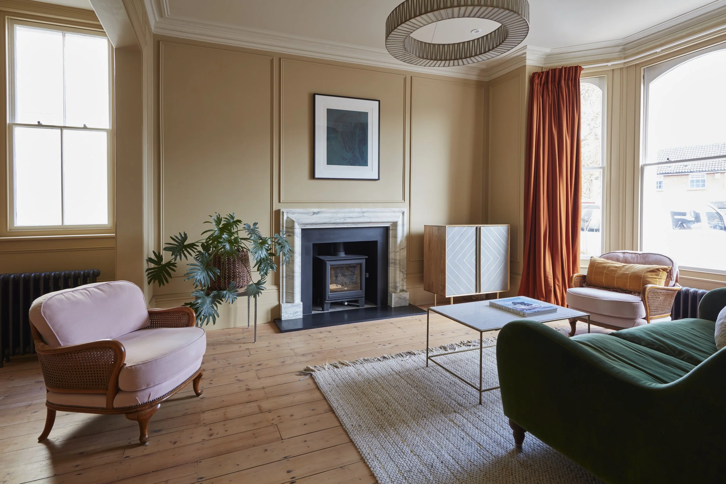A cozy living room with beige walls, a marble fireplace, and large windows with orange curtains. There are vintage-style sofas and chairs, a coffee table with books, a patterned rug, a potted plant, and wall art.