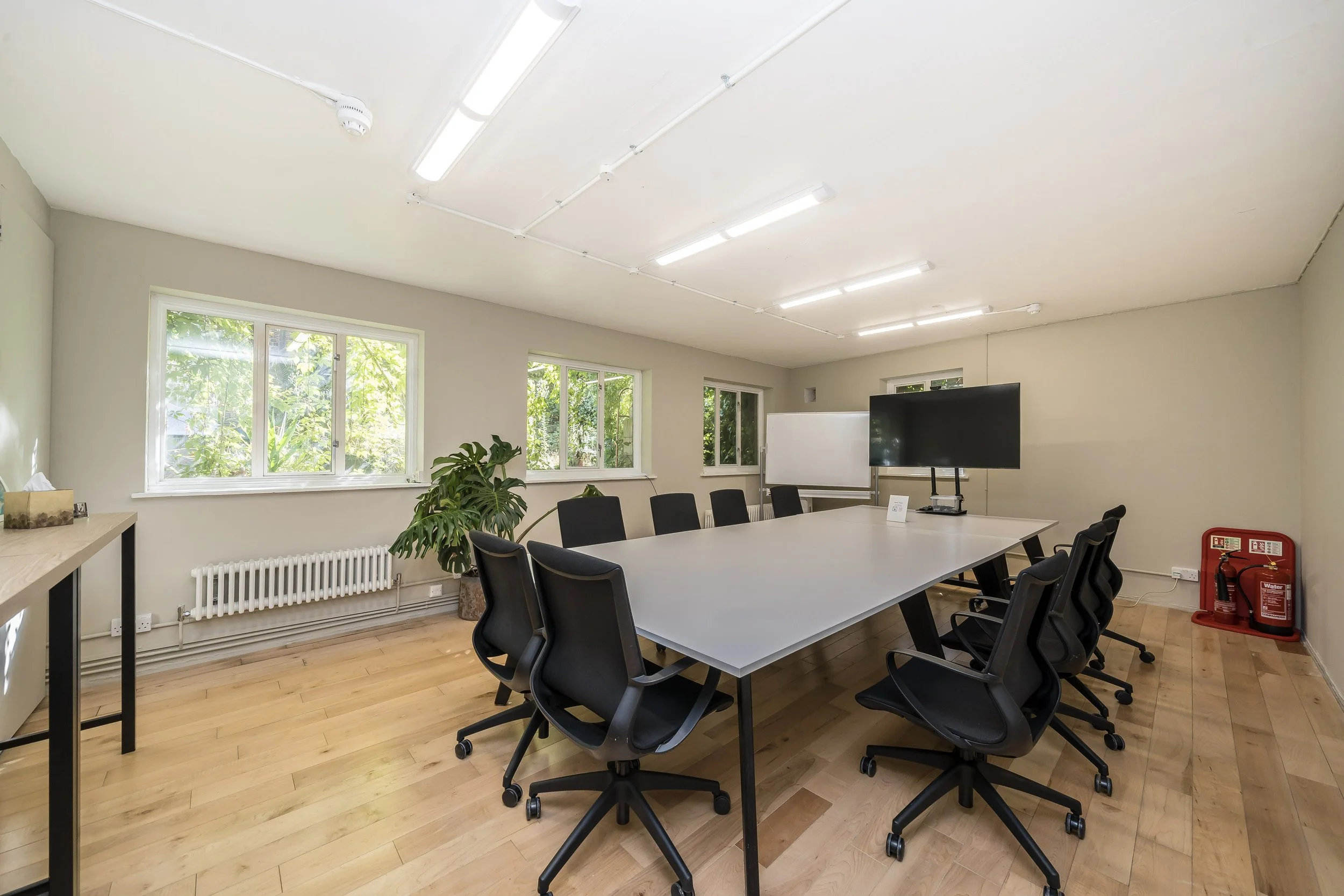 Empty conference room with a long table, black chairs, windows with greenery outside, whiteboad, large screen, and fire extinguisher.