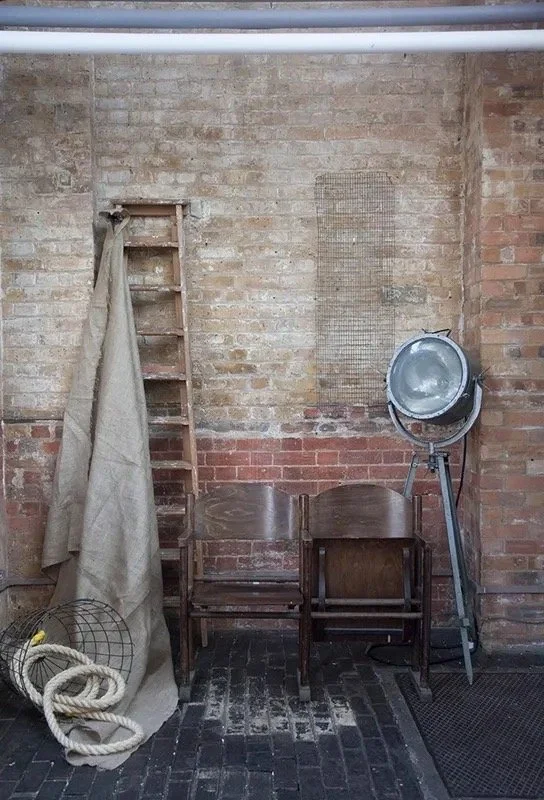 Interior of an industrial space with exposed brick wall, two wooden chairs, a hairdressing hood dryer, a ladder draped with a cloth, and a coiled rope and wire basket on the floor.