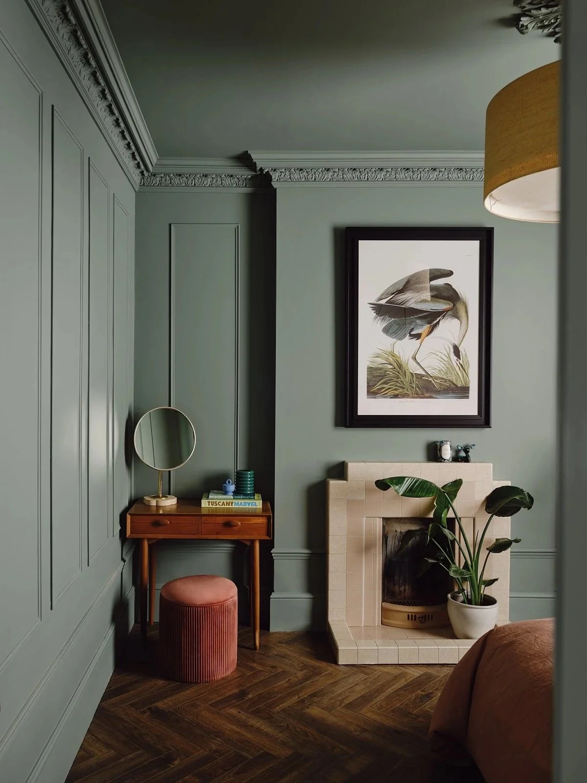 A cozy corner of a living room with gray green paneled walls, a fireplace with a beige tiled mantle, a potted plant, a small wooden vanity with a mirror, a pink velvet stool, and framed artwork of a heron hanging above the fireplace.