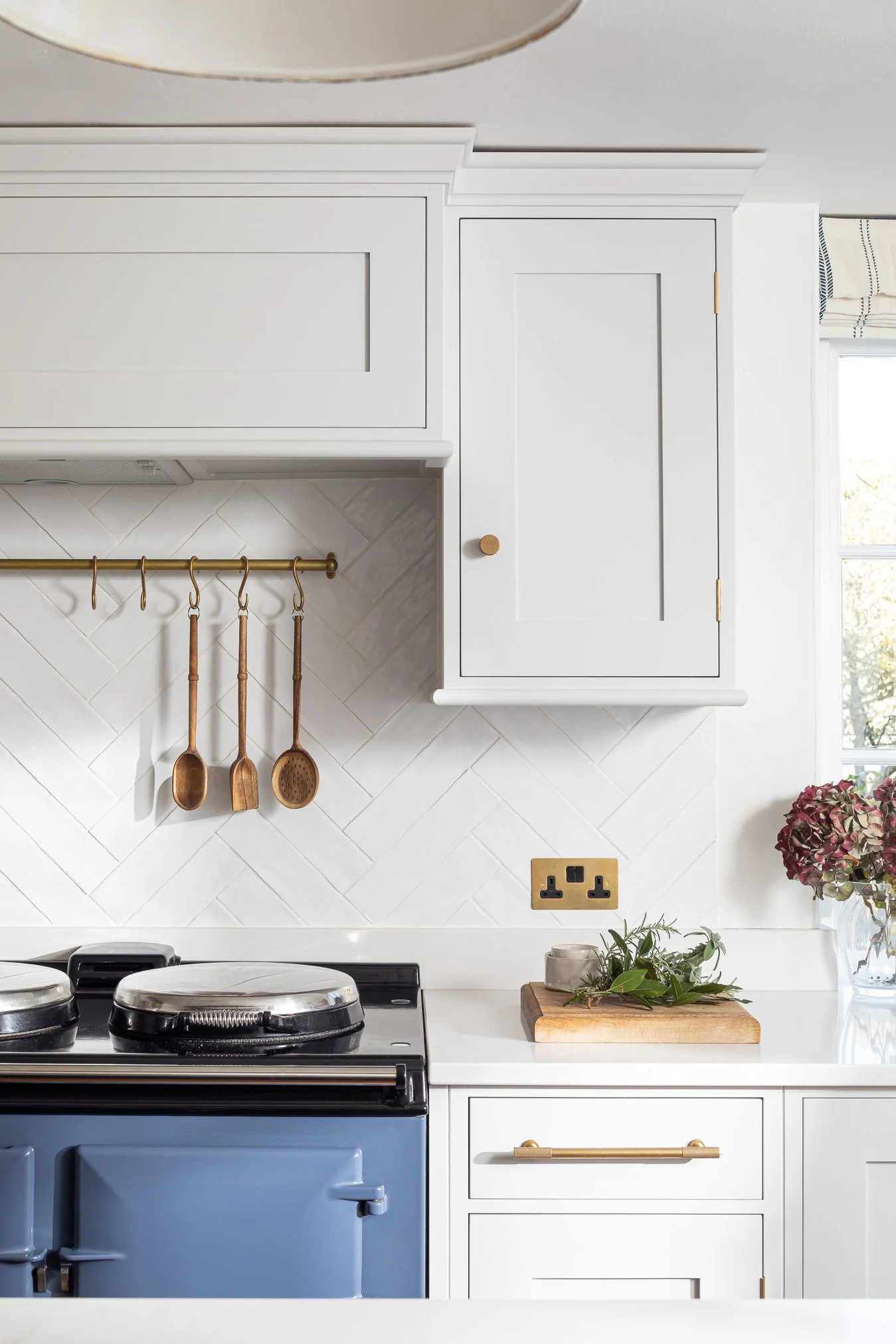 A kitchen countertop with a black cast iron stove, gold kitchen utensils hanging on a rail, a bouquet of flowers, a cutting board with herbs, and a white cabinet with gold handles. There is a window with a view outside.