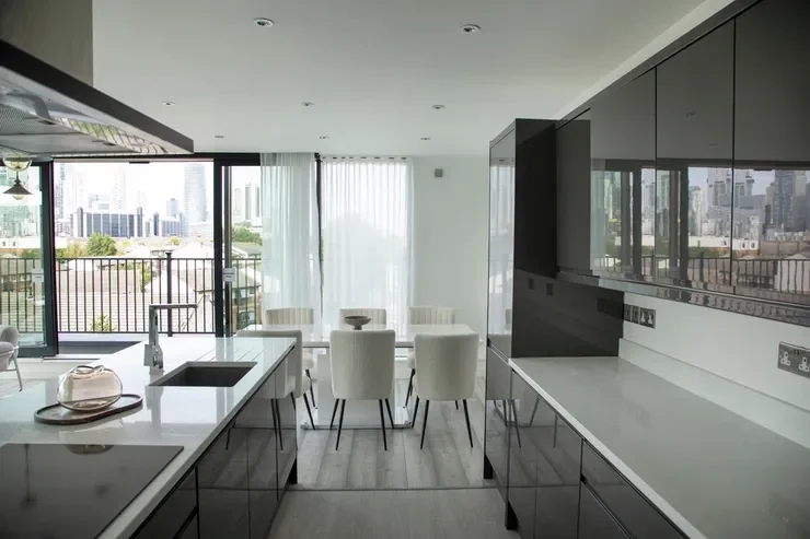 Modern kitchen and dining area with white countertops, black cabinets, and a set of six white chairs around a round dining table, with large windows and city view outside.