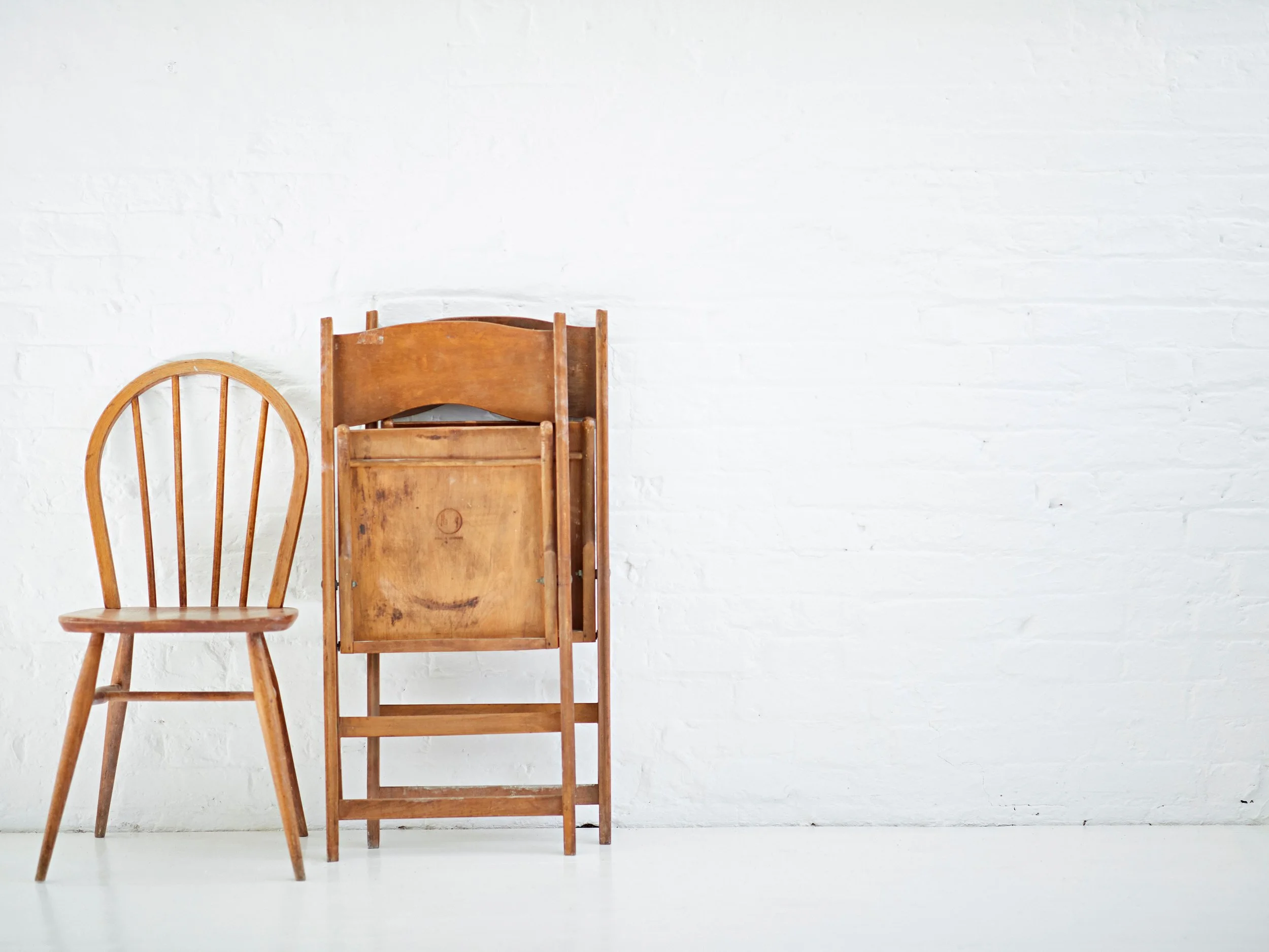 Two wooden chairs, one with a traditional wooden frame and the other a folding chair, placed side by side against a white brick wall.