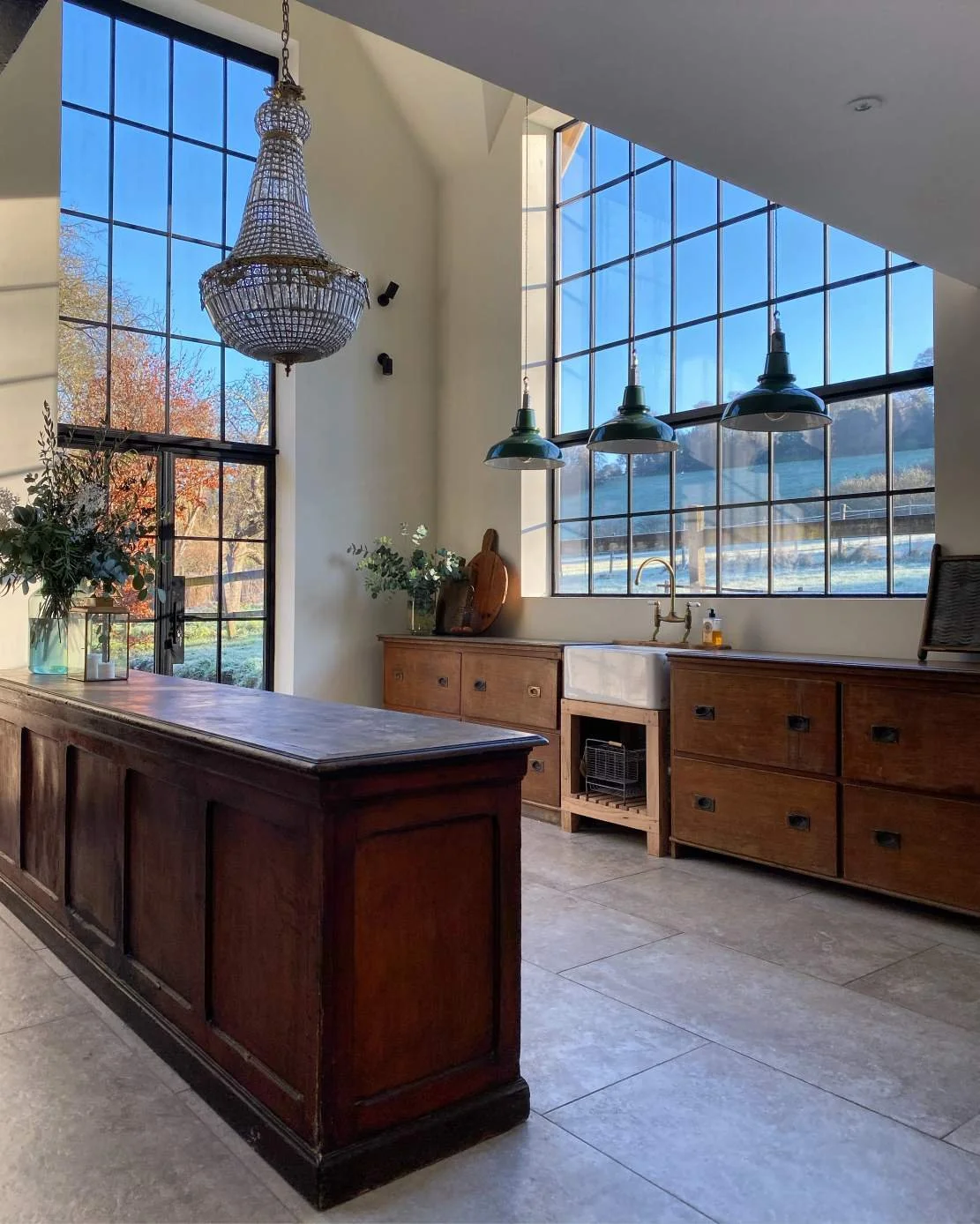 A bright kitchen with large grid windows, wooden cabinets, a farmhouse sink, green pendant lights, a chandelier, and a view of fall foliage outside.