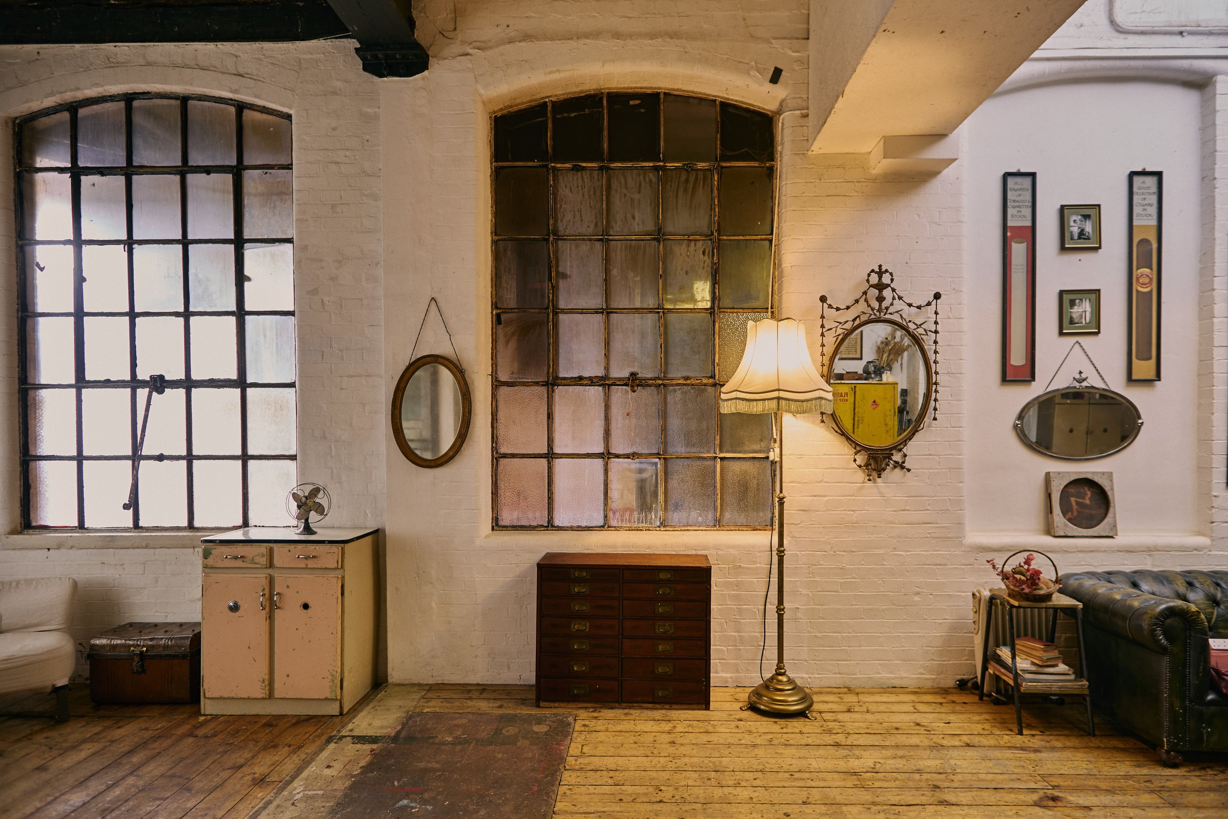 Interior of a vintage-style room with large industrial windows, a white brick wall, a standing floor lamp with a lampshade, a small wooden cabinet with drawers, and a black leather sofa. Decor includes mirrors, framed pictures, and a small side table