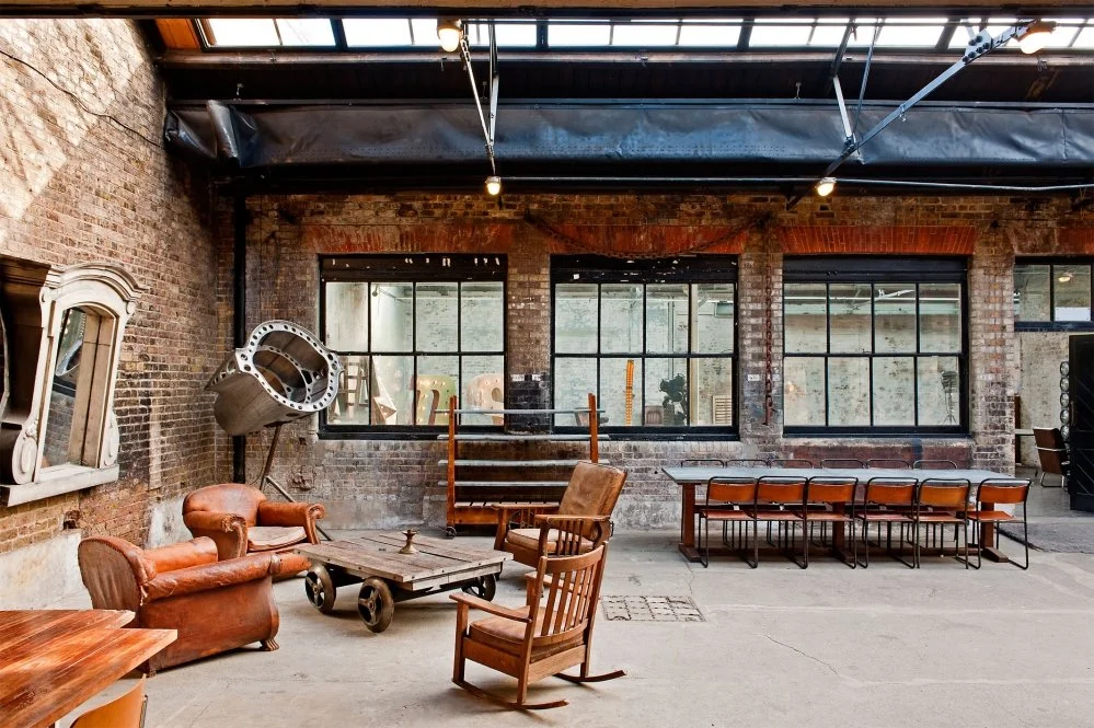 Industrial-style loft with exposed brick walls, large windows, vintage furniture including a leather armchair, wooden chairs and tables, and an artwork installation.
