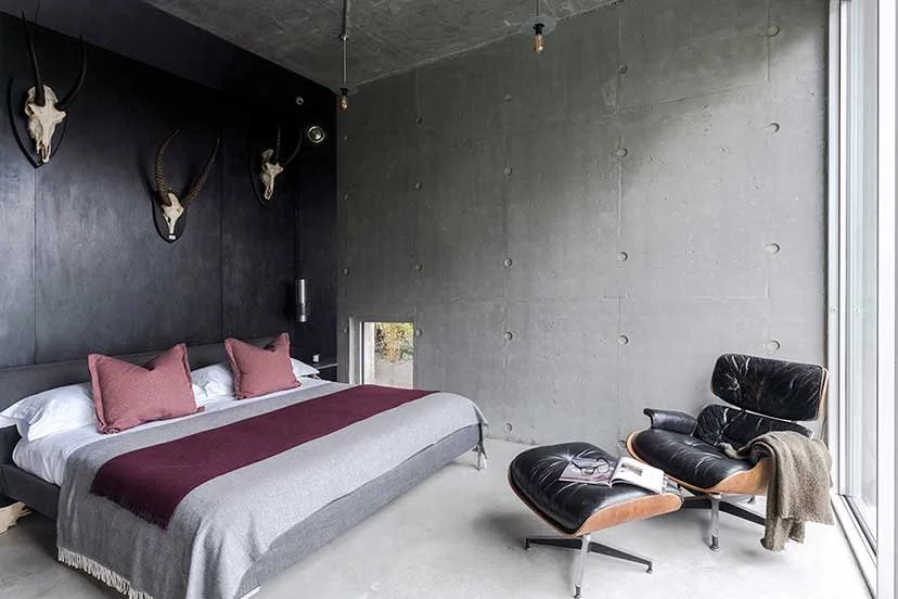 Modern bedroom with concrete walls, mounted animal skulls, large bed with pink and gray bedding, black leather lounge chair and ottoman with a blanket, large window