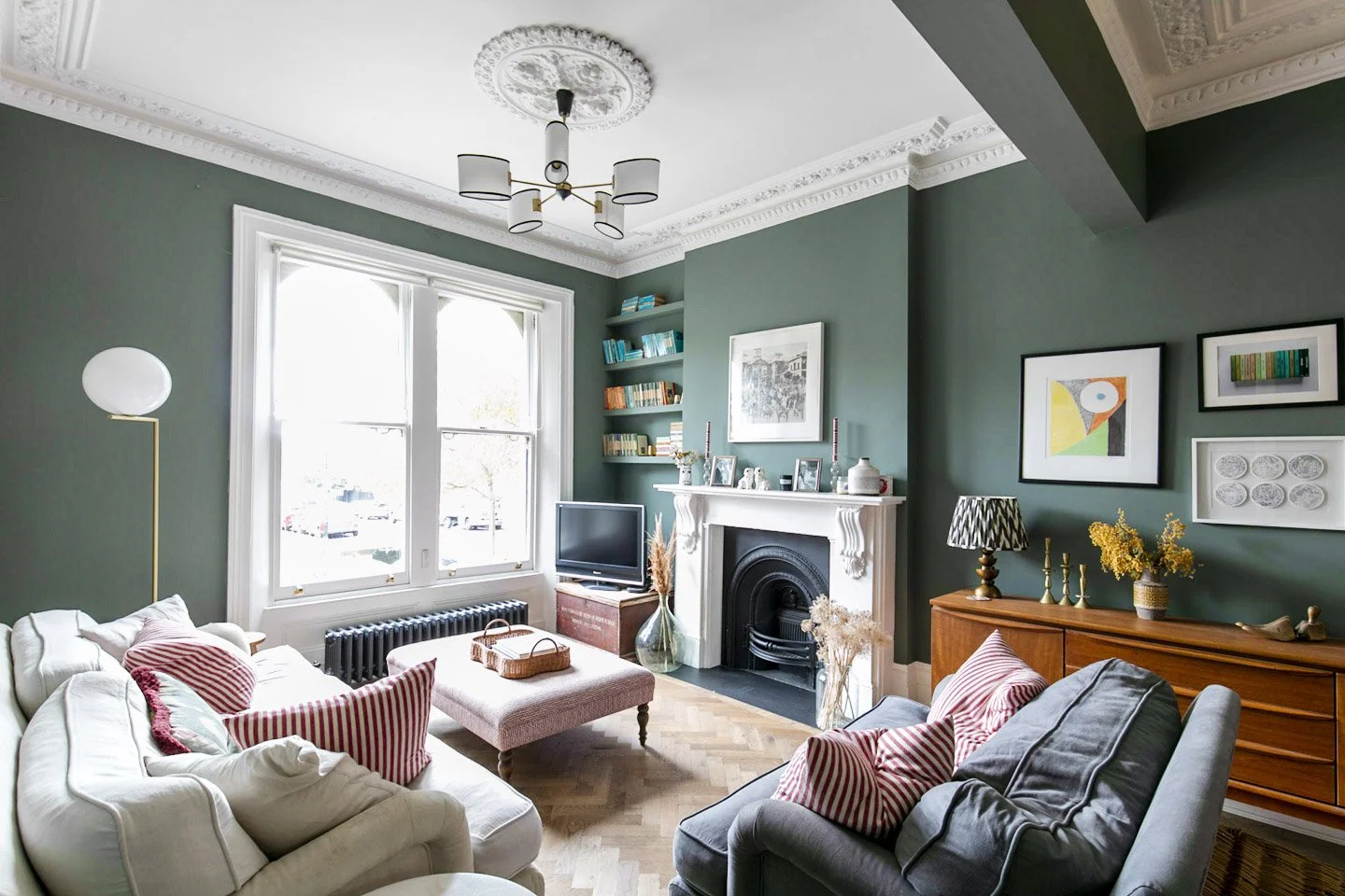 Living room with green walls, large window, white sofa with red striped pillows, a pink ottoman, a grey seating area, a wooden sideboard with yellow flowers, wall art, a black fireplace, a nearby TV, and a coat rack.