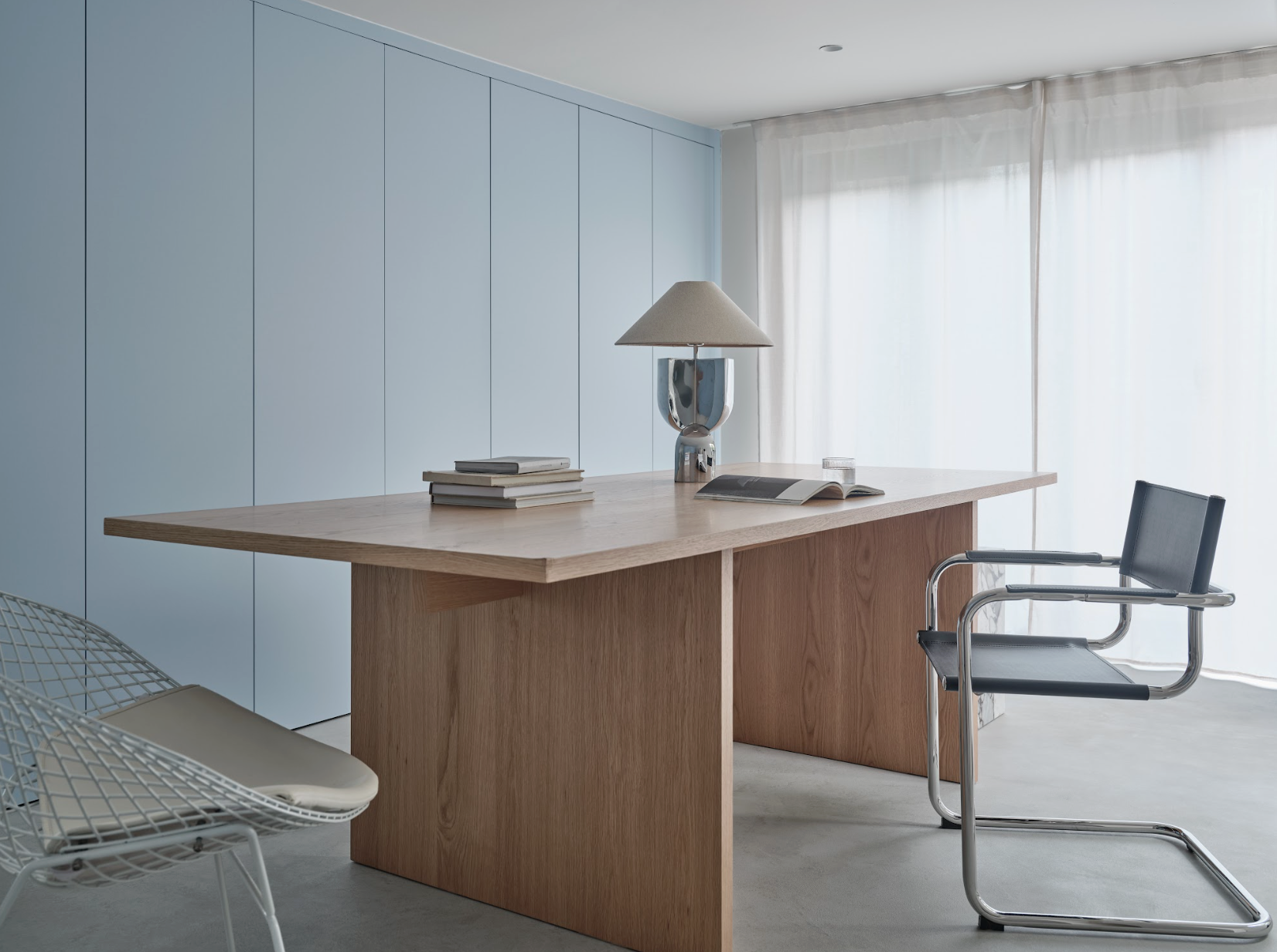 Modern minimalist home office with wooden desk, black chair, white wire chair, blue wall cabinets, and sheer curtains.