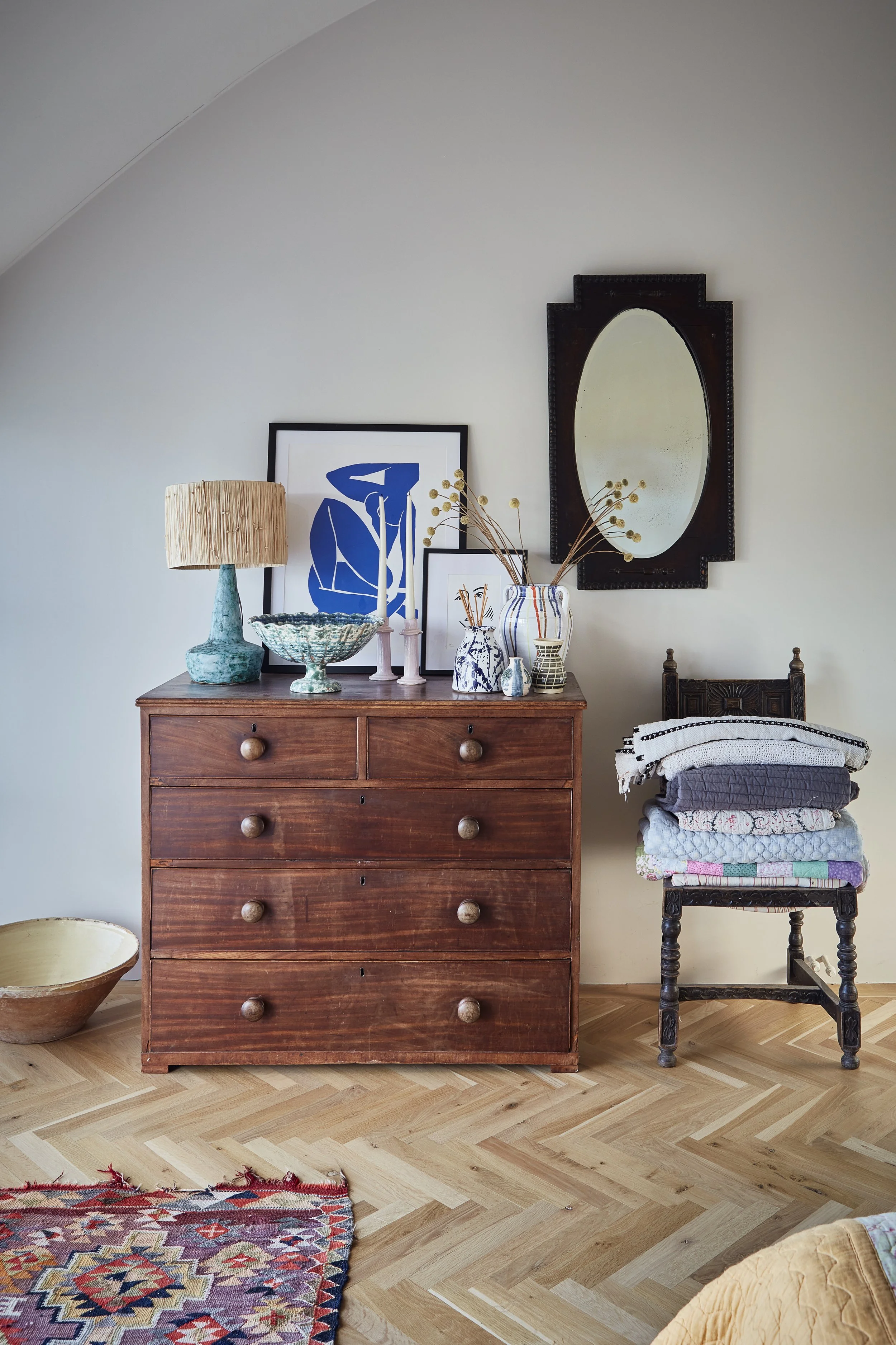 Various decorative objects on a wooden dresser, including a beige lamp, framed blue abstract art, vases, and dried plants, against a white wall with a round mirror. To the right, a chair supports a stack of folded textiles, and part of a patterned ru