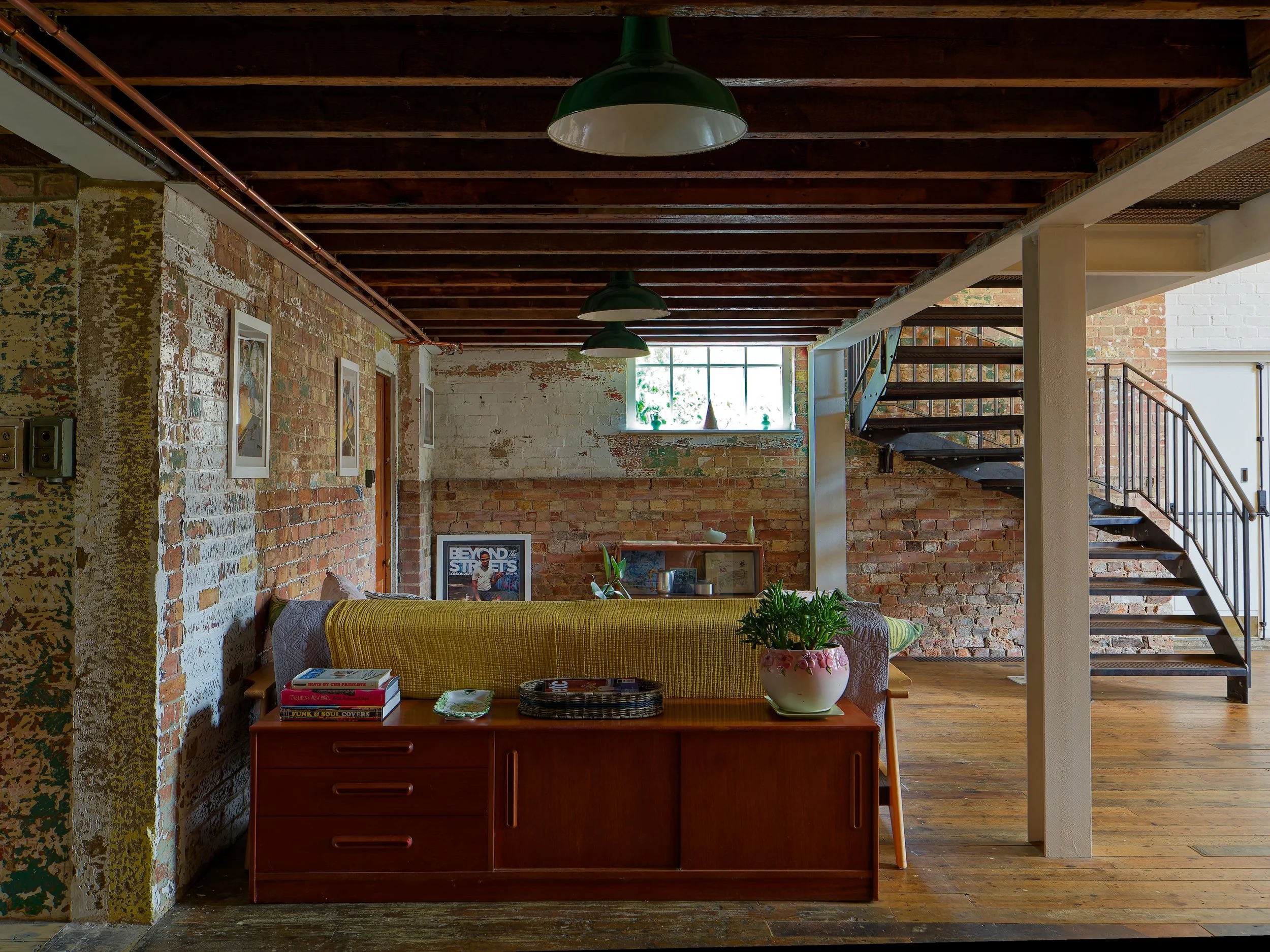Interior view of a loft-style living space with exposed brick walls, a wooden floor, a staircase, and a window letting in natural light. There is a wooden sideboard with books and a potted plant, and framed artwork on the walls.
