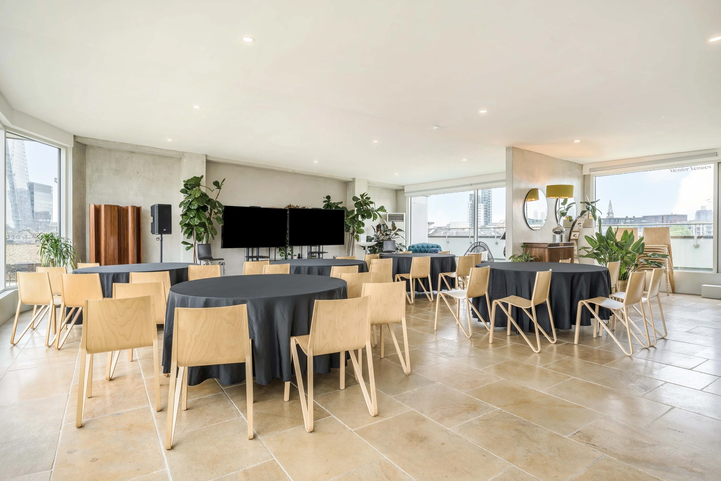 Conference or event room with round tables covered in black tablecloths, surrounded by light wood chairs, large windows, green plants, a TV, and a mirror on the wall.