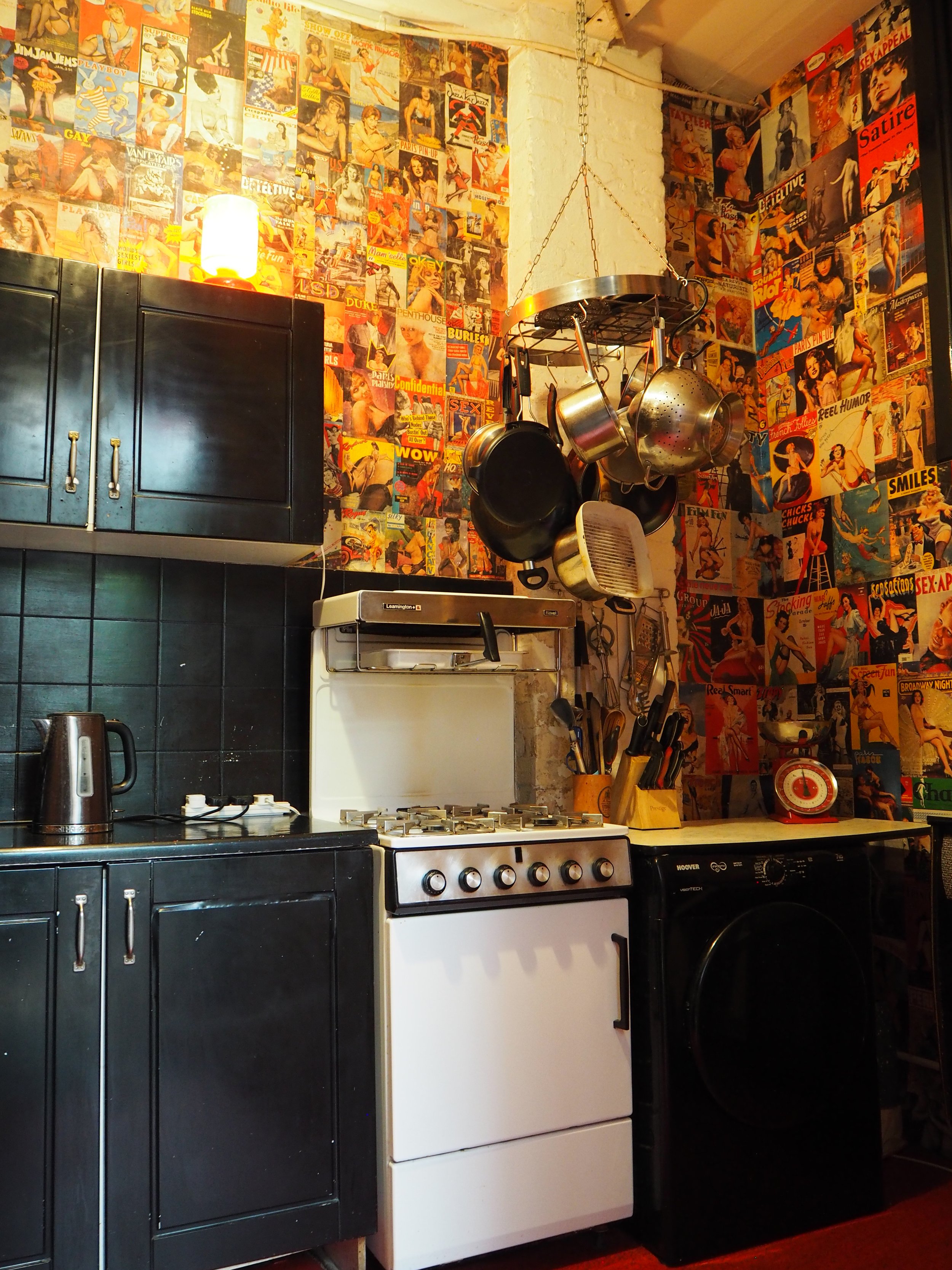 A kitchen corner with black cabinets, a white stove, and a black washing machine. Above, a hanging pot rack holds several pots and pans. The wall is decorated with colorful vintage magazine covers. There is a timer and some kitchen utensils on the co