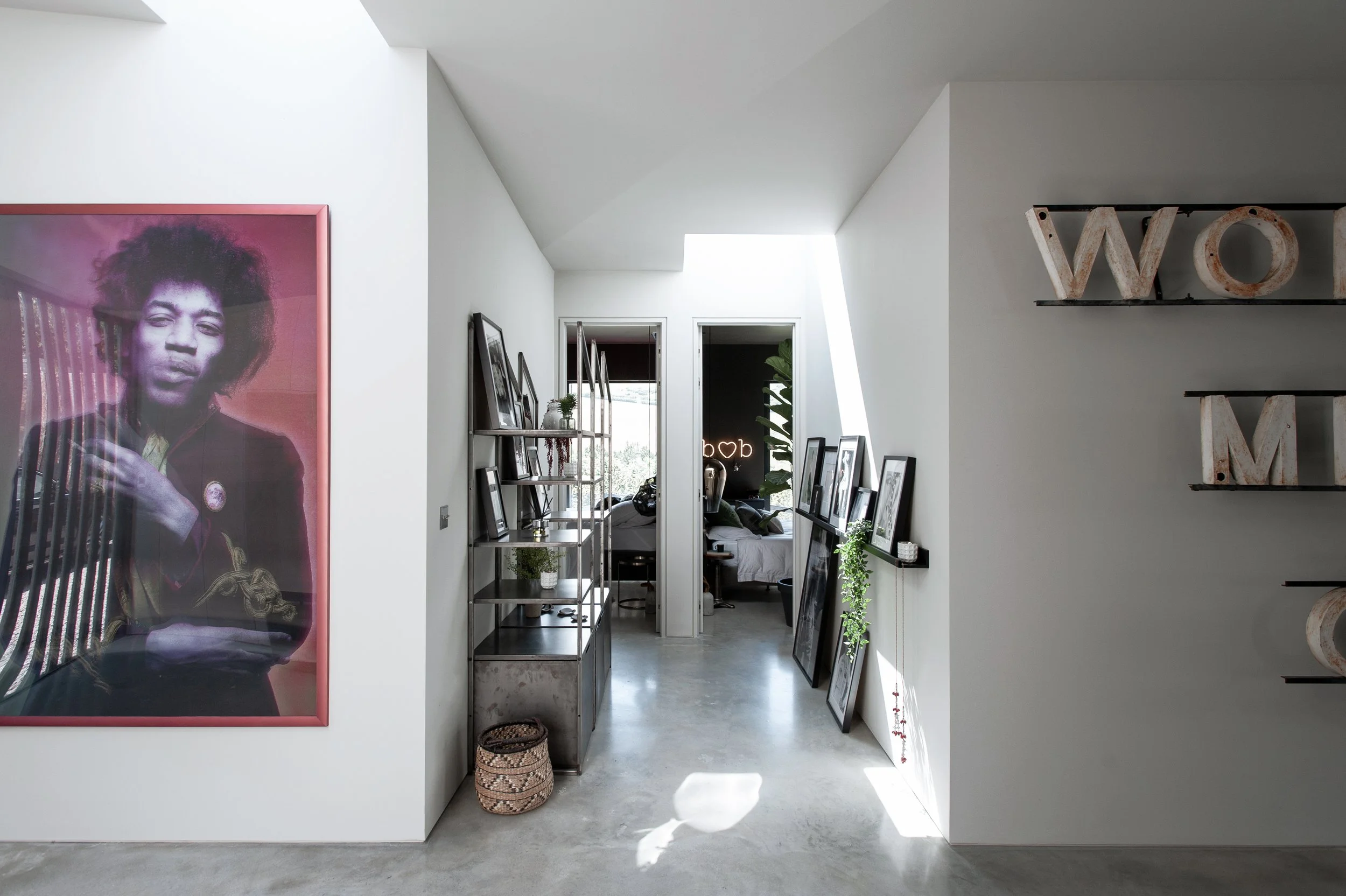 Modern interior hallway with black and white framed photographs leaning against wall, a metal shelf with small potted plants, and natural light flooding through a skylight. A large portrait of a woman with curly hair is on the left, and shelves with 