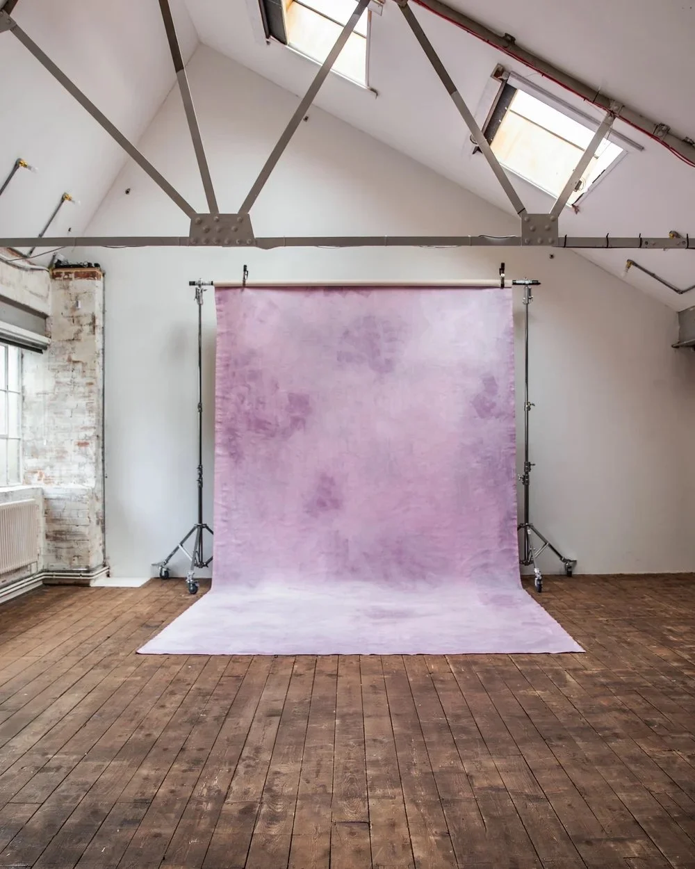 An indoor photo of a photography studio with a purple backdrop on a stand, wooden floor, white walls, two windows on the ceiling, and visible metal ceiling beams.