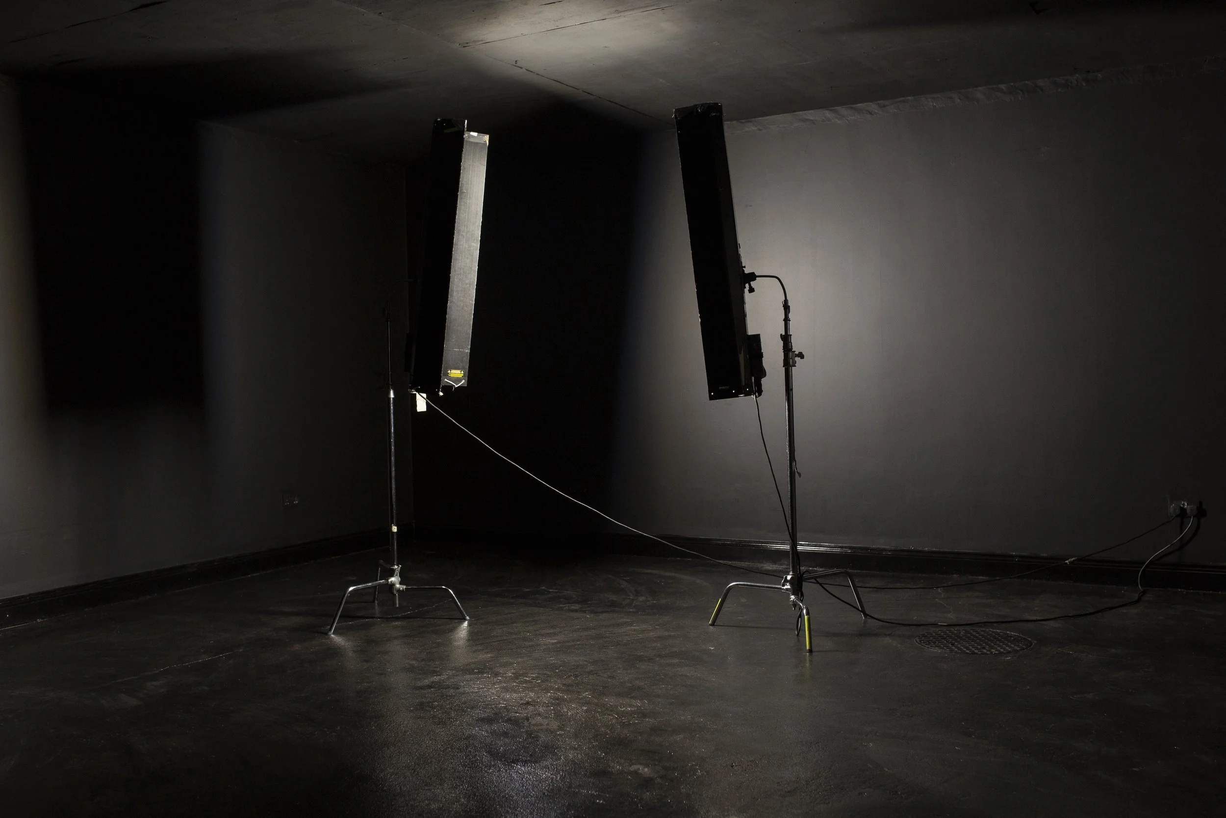 Dark, empty room with two standing studio lights on tripods, turned off, connected by a cable, and a drain cover on the floor.