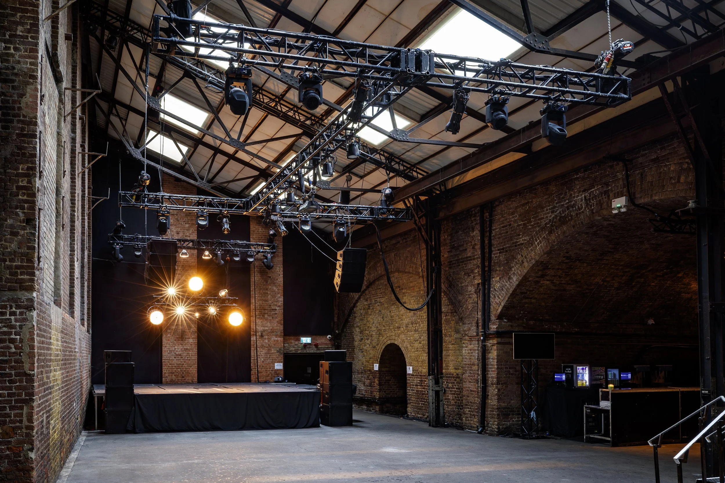 An empty event venue with brick walls, a stage, and stage lighting and sound equipment, including spotlights and large speakers.