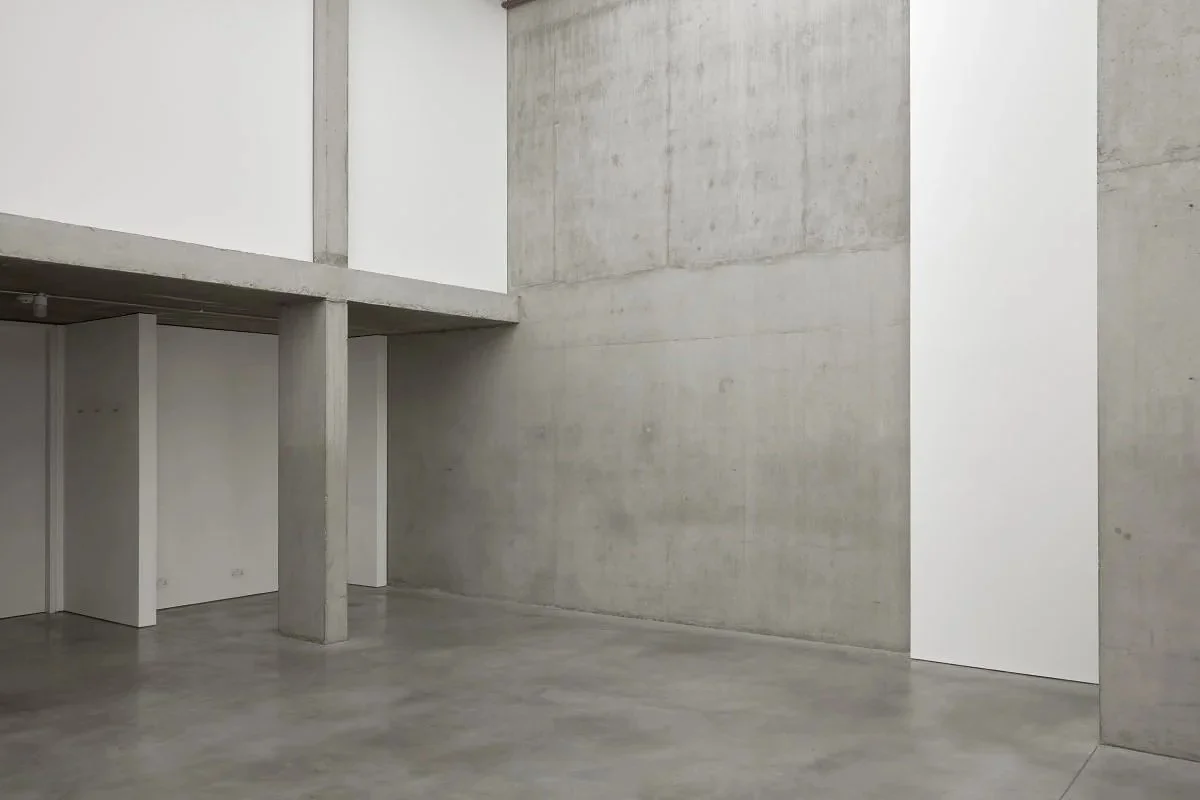 Empty industrial-style room with concrete walls, floor, and ceiling. Some white panels or partitions are seen, with one opposite corner.