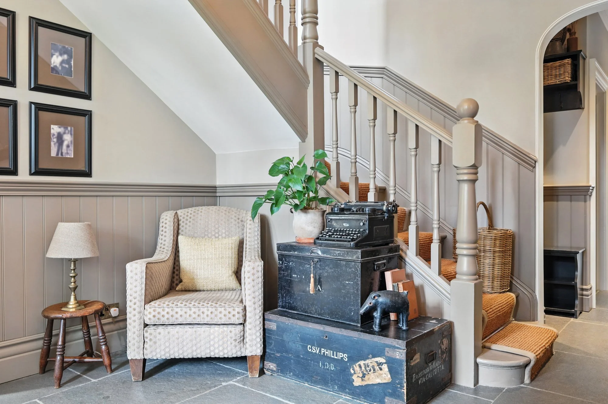 Living room corner with beige armchair, small wooden stool with lamp, potted plant, vintage typewriter on stacked black trunks, staircase with white banister, framed photographs on wall, wicker basket, and decorative items in an archway.