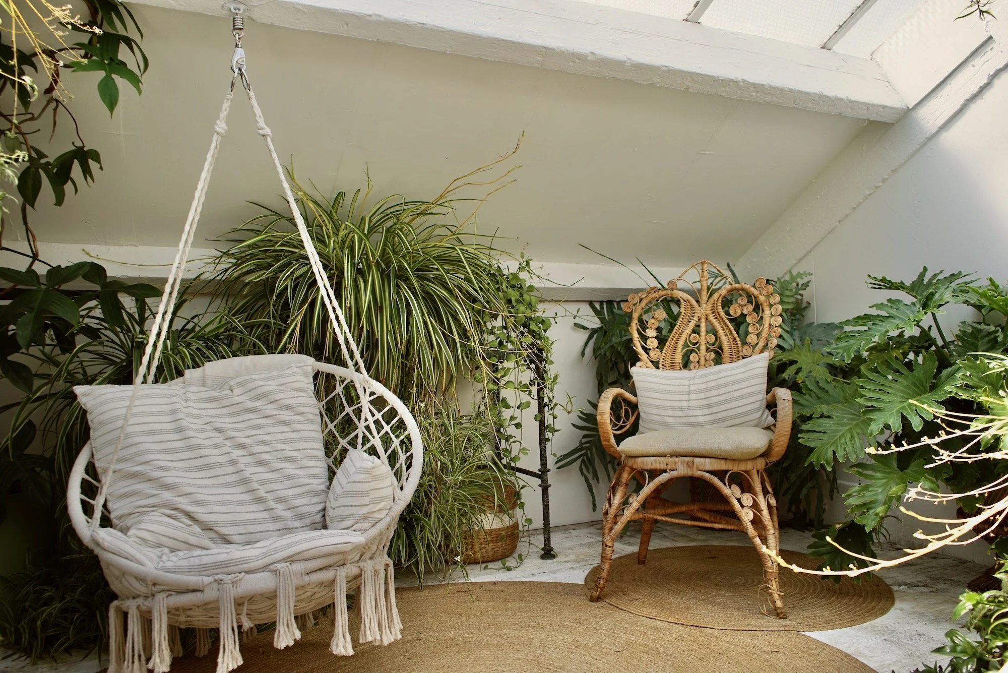 Indoor cozy porch with white hanging chair and wooden armchair surrounded by green plants and natural fiber rugs.