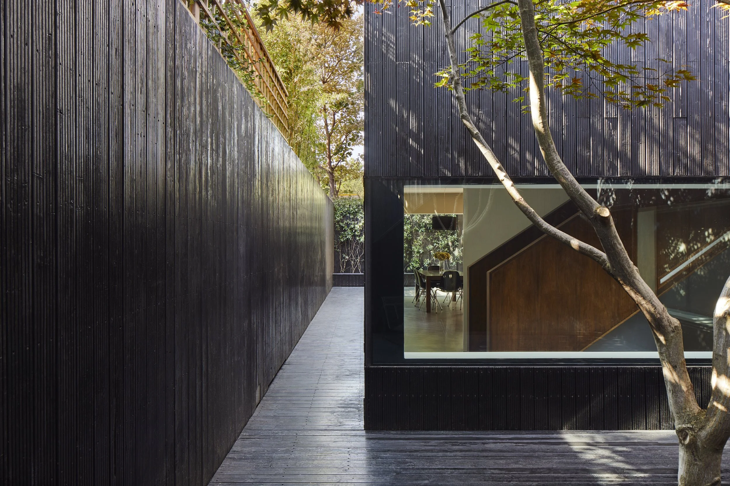 Modern building with black exterior walls, a large window showing an interior dining area, and a tree with branches extending in front.
