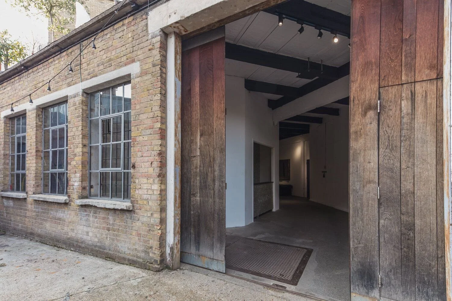 Exterior view of a building with brick walls and large grid windows, featuring a wide open wooden door leading into a modern indoor space with visible ceiling beams.