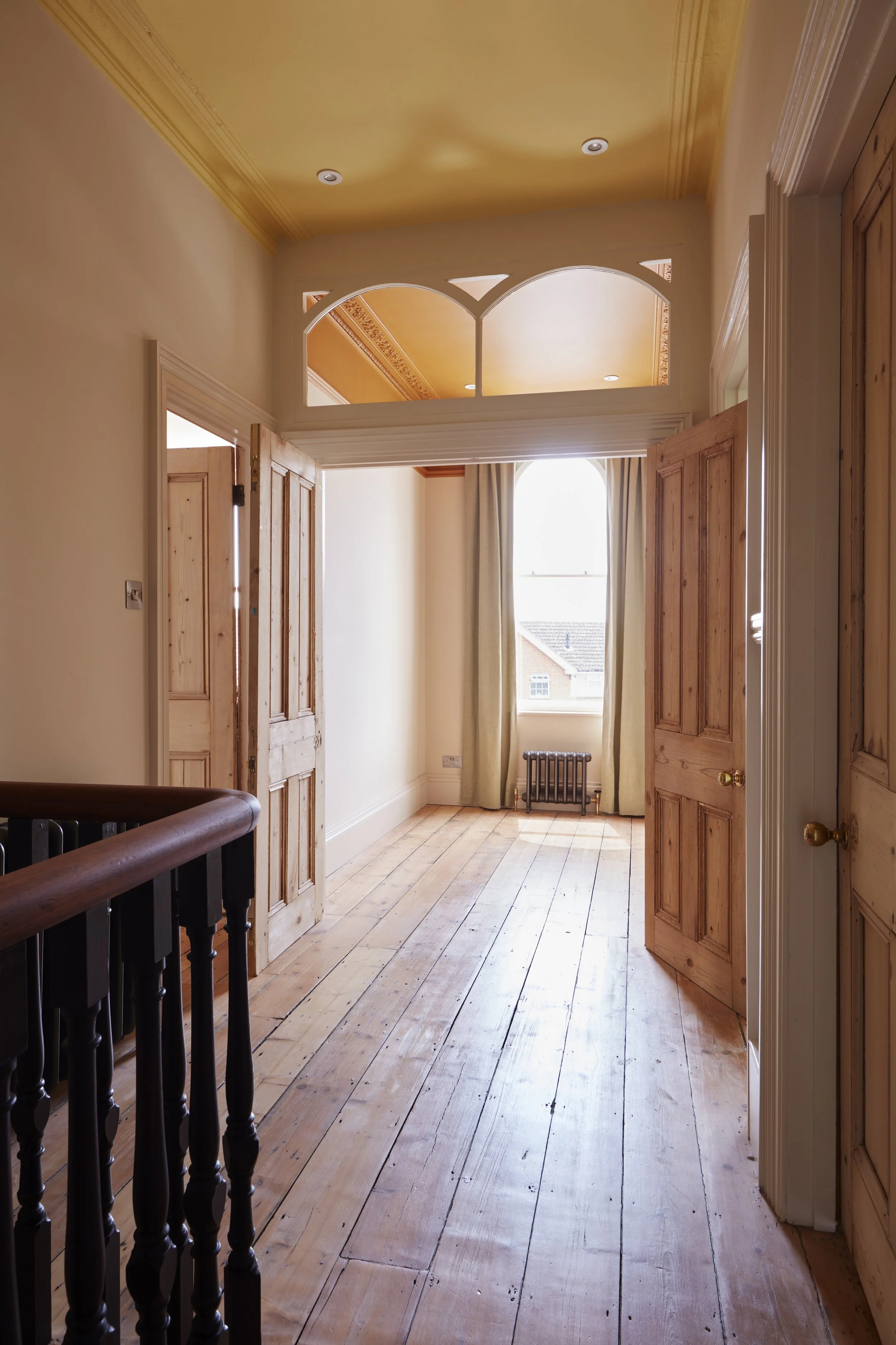 Empty hallway with wood flooring and open wooden doors, window with beige curtains, and ceiling with recessed lighting.