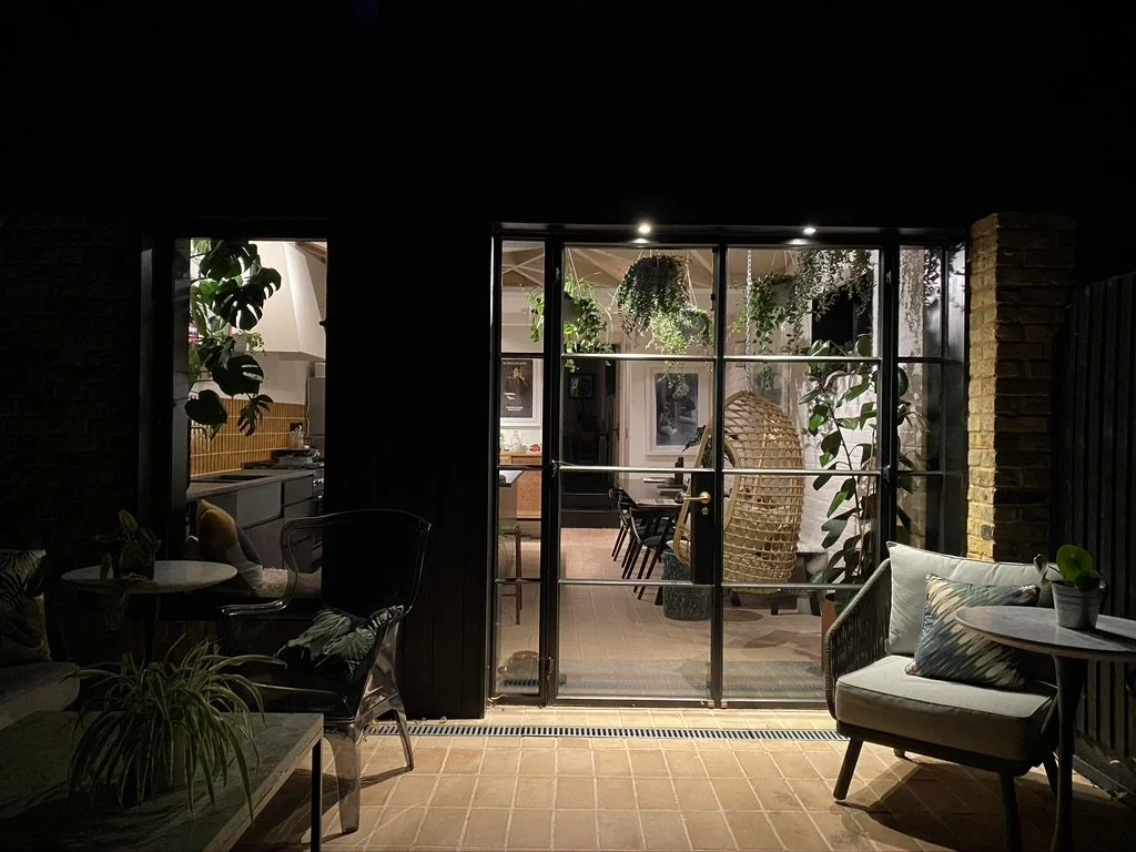Interior view of a cozy room looking through glass sliding doors to an outdoor patio space filled with hanging plants and outdoor chairs.