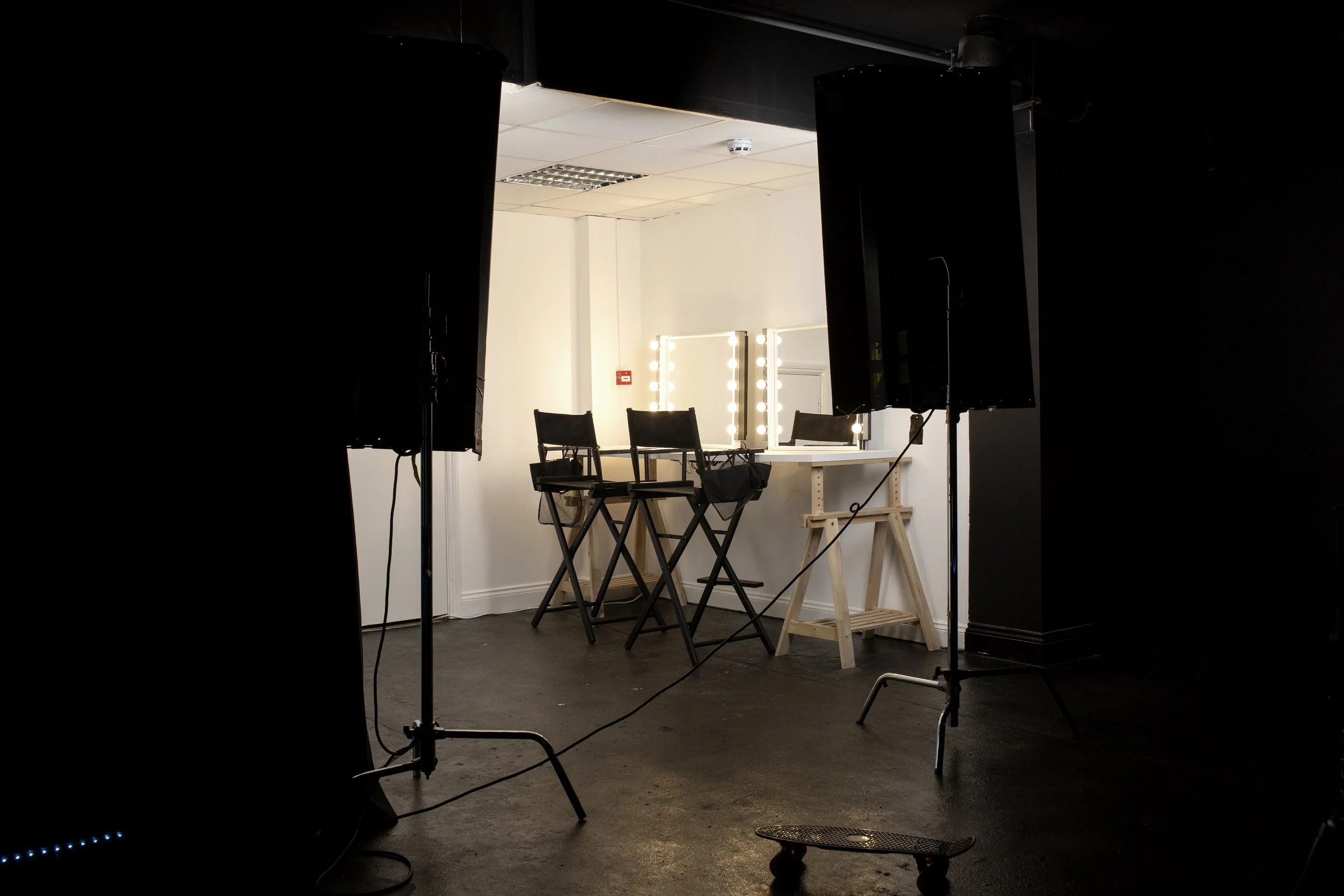 A photography studio setup featuring a white wall with a lit makeup mirror, three black chairs on a wooden table, and studio lights on stands.