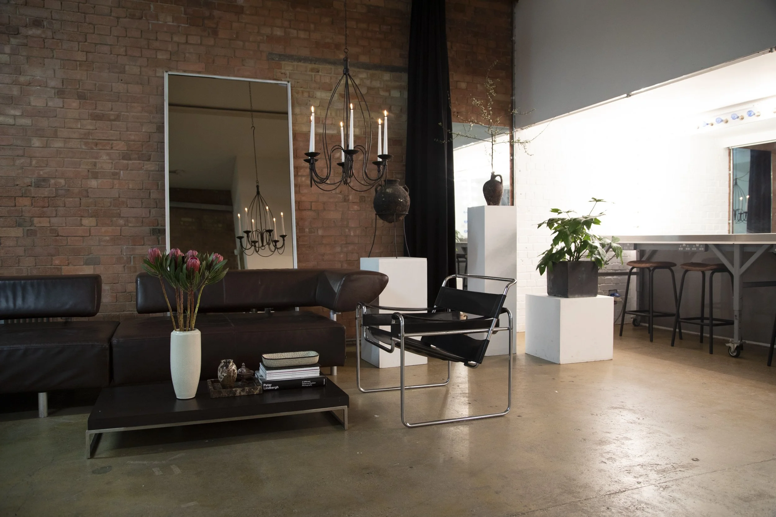 Modern living room with brick wall, leather sofa, coffee table with books and decorative items, vases with flowers and plants, large mirror, chandelier, and bar seating area.