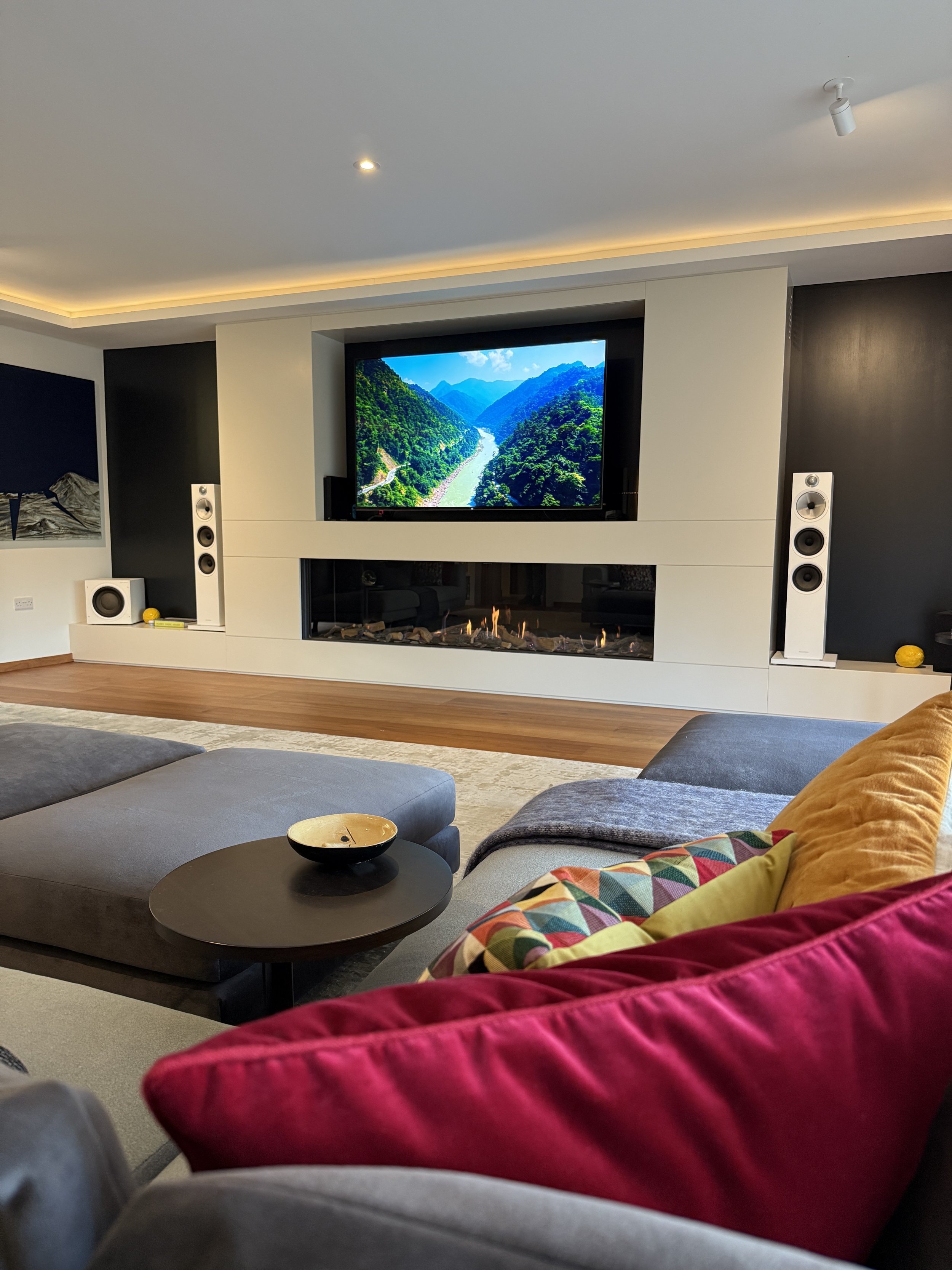 Living room with a large flat-screen TV showing mountains and a river, surrounded by modern speakers and a fireplace, with colorful cushions on a sofa and a small side table.