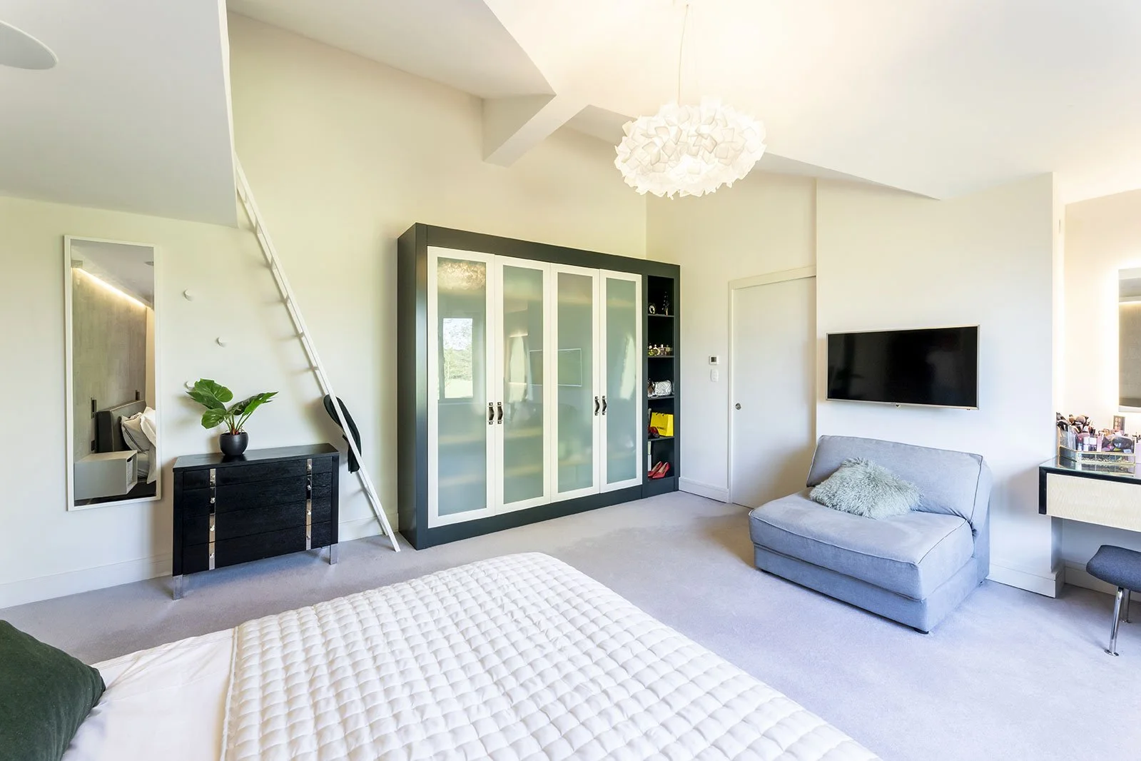 Modern bedroom with white walls, a bed with white quilt, a gray chair with a pillow, a black dresser with a plant, a large mirror, a black and white wardrobe, a flat-screen TV, and a small vanity or desk area with makeup items.