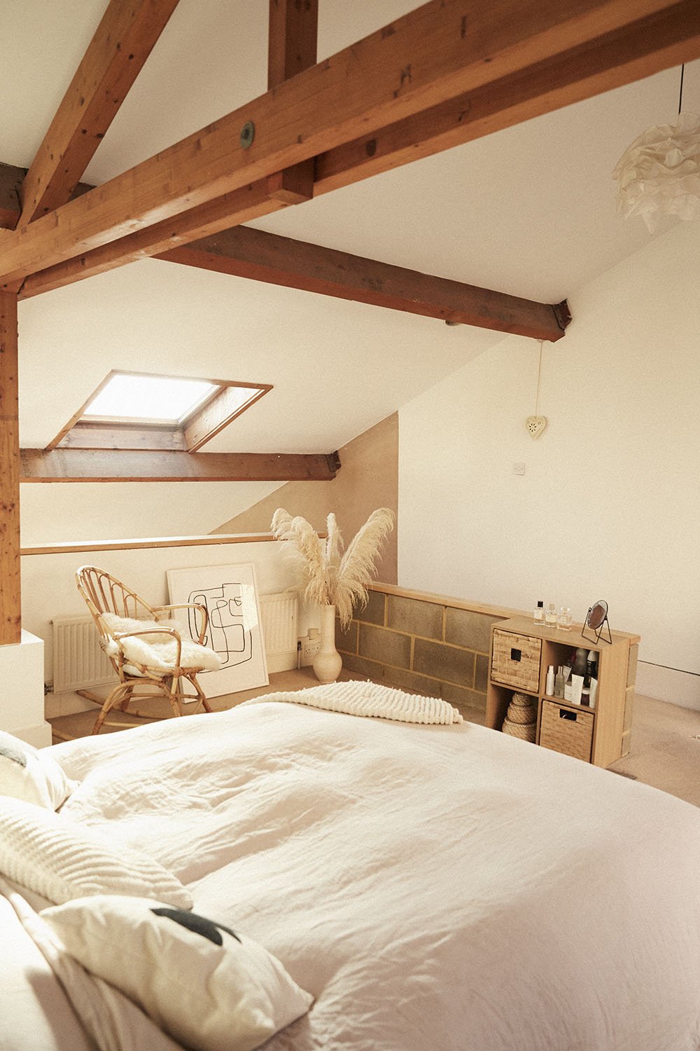 Bright bedroom with sloped ceiling, wooden beams, a skylight, and cozy decor including a bed, a wicker chair, a framed artwork, a large vase with pampas grass, a small wooden storage unit, and hanging heart decoration.