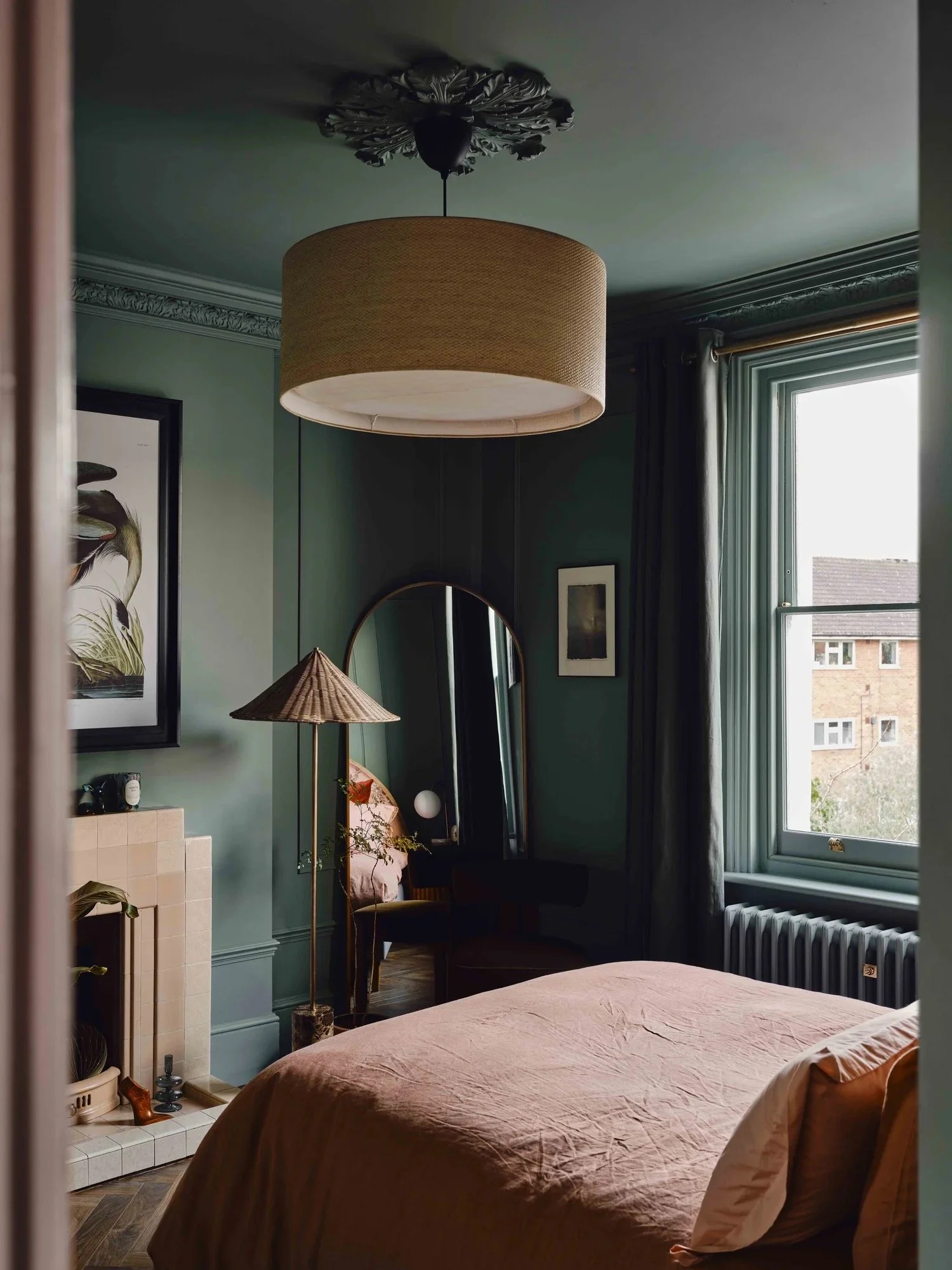 A cozy bedroom with green walls, a large window with curtains, a bed with a beige bedspread, a tall mirror, a lamp with a straw shade, and artwork on the walls.