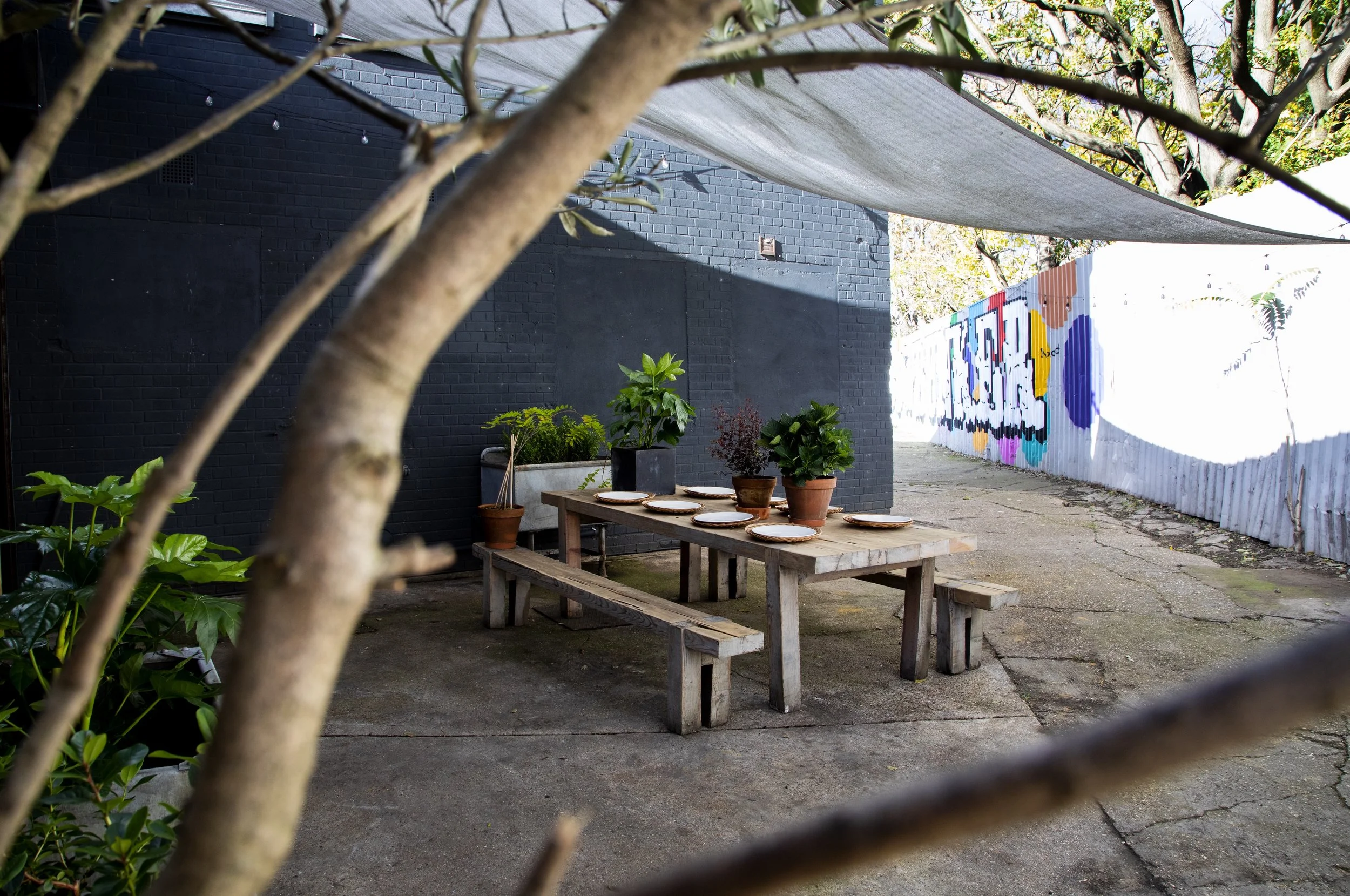 Outdoor patio scene with a weathered wooden table and benches, decorated with several potted plants and plates, set against a black brick wall, with a painted mural on a corrugated metal fence in the background, and a fabric shade overhead.