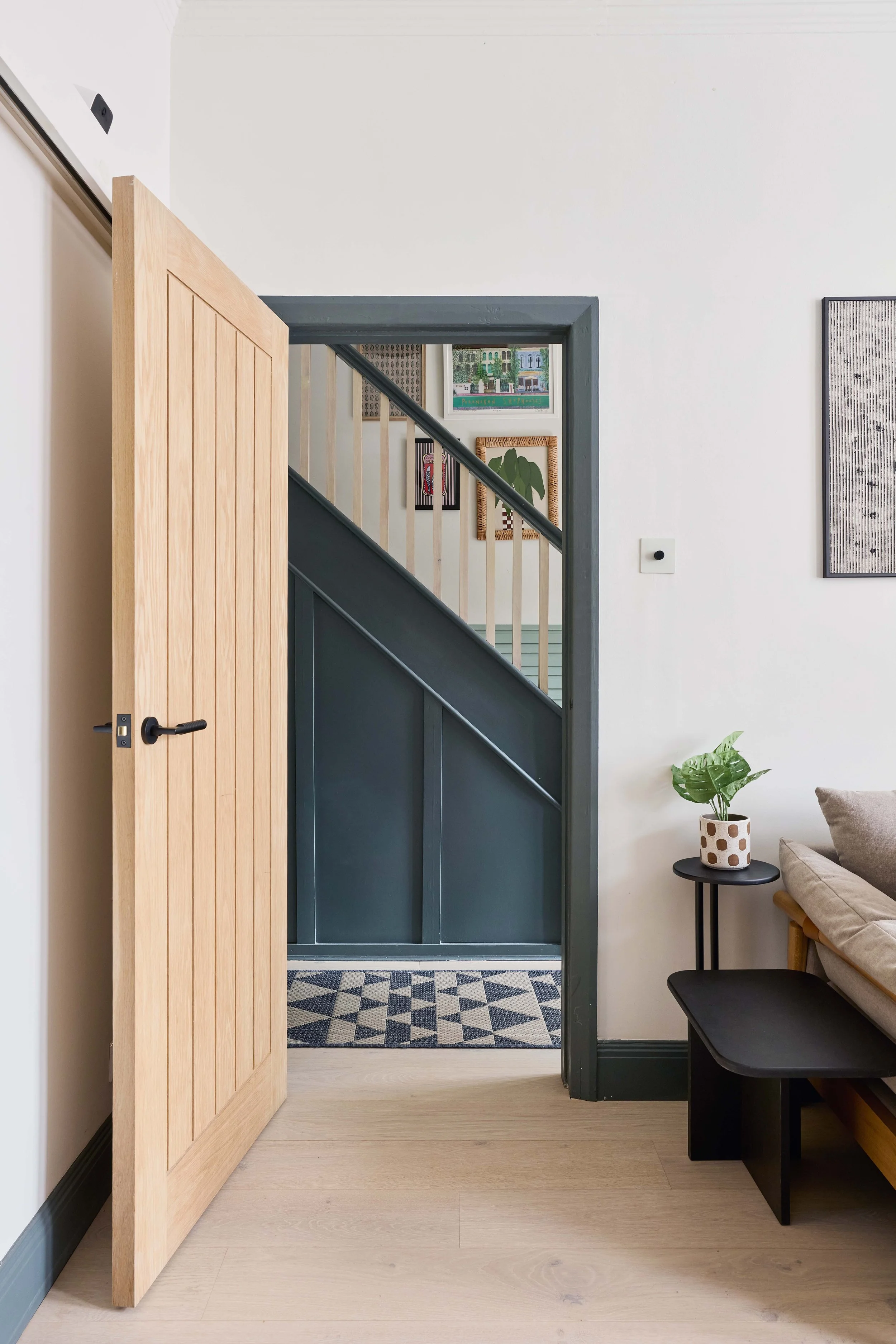 Interior view of a living space with a wooden door partially open, a small black side table with a potted plant, beige couch with a pillow, and wall art. Visible staircase through the doorway.