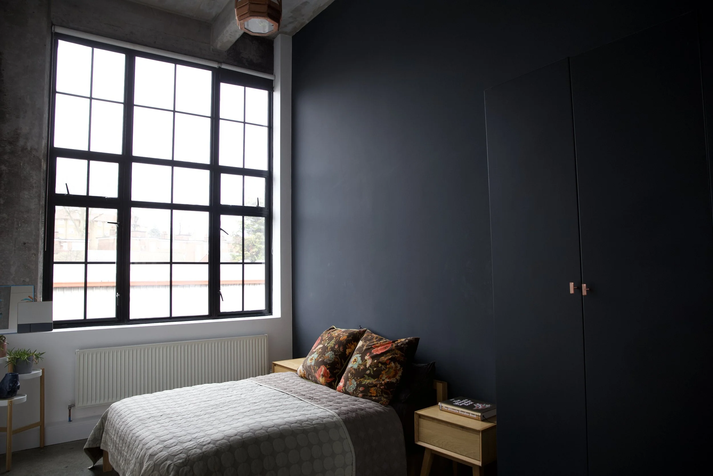 Bedroom with a large grid window, dark blue accent wall, bed with floral pillows, bedside tables with books, and a closet.