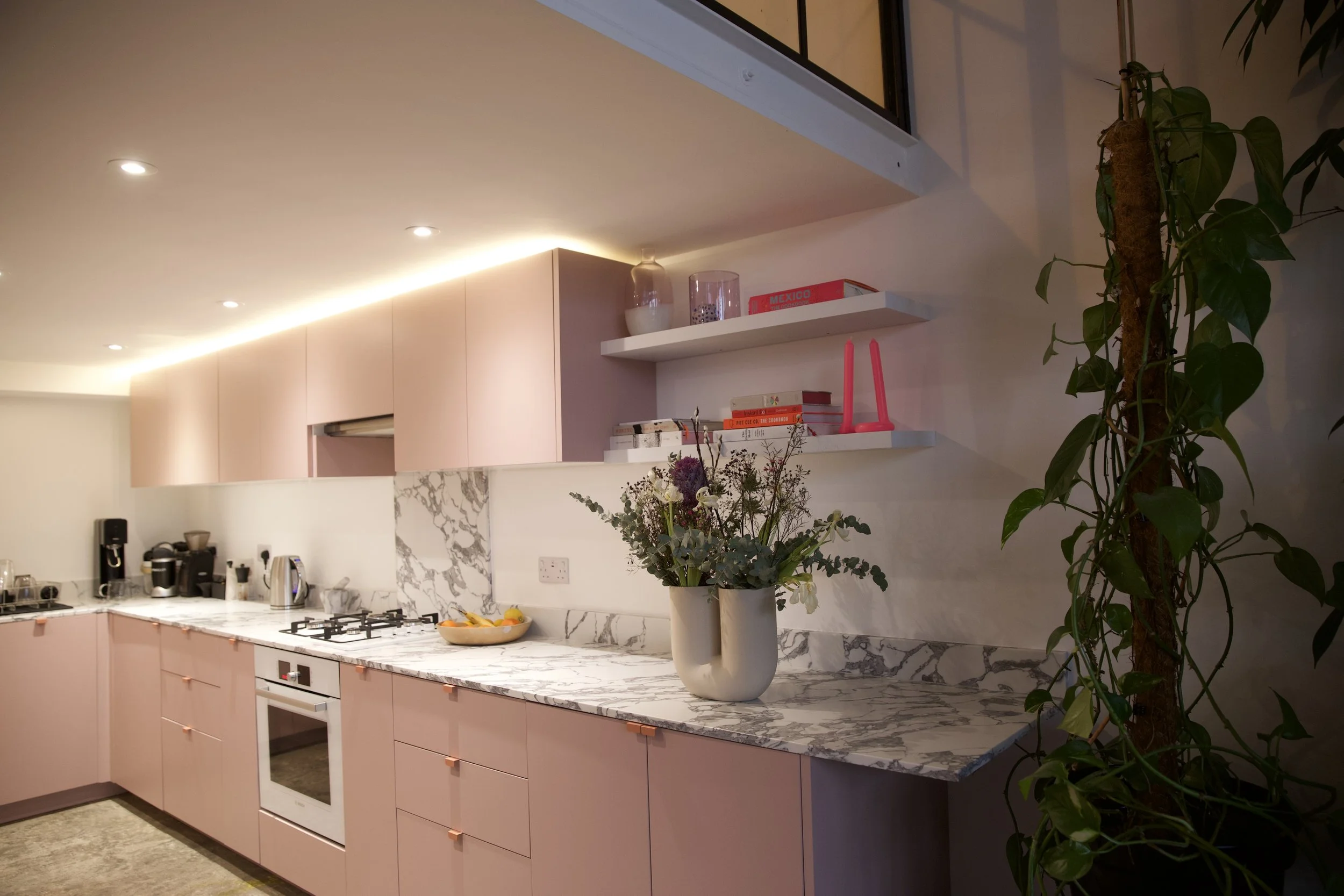 Modern kitchen with pink cabinets, marble countertops, the open shelving with books and pink candle holders, a vase with flowers, and a large green climbing plant.
