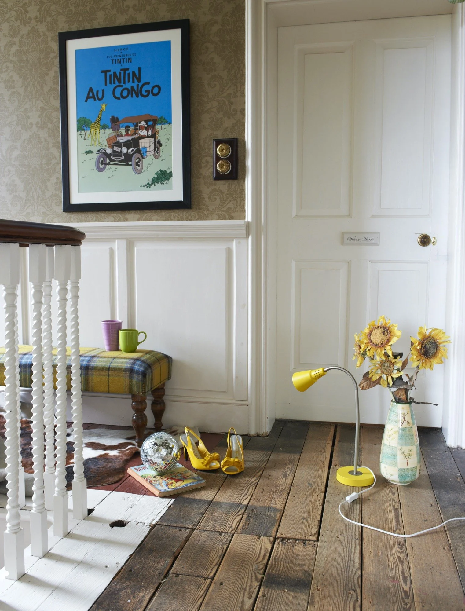 Interior hallway with wooden floor, a framed Tintin poster on the wall, a small bench with colorful mugs, a vase with yellow sunflowers, yellow shoes, a disco ball, a magazine, a yellow desk lamp, and a white door.