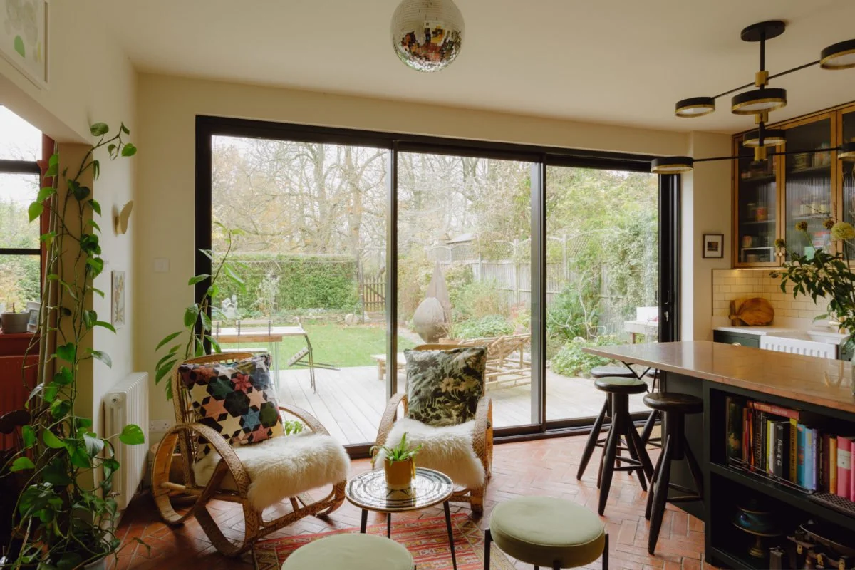 Living room with large sliding glass doors opening to a backyard, featuring two armchairs with furry throws, a small round table, and barstools near a kitchen island.
