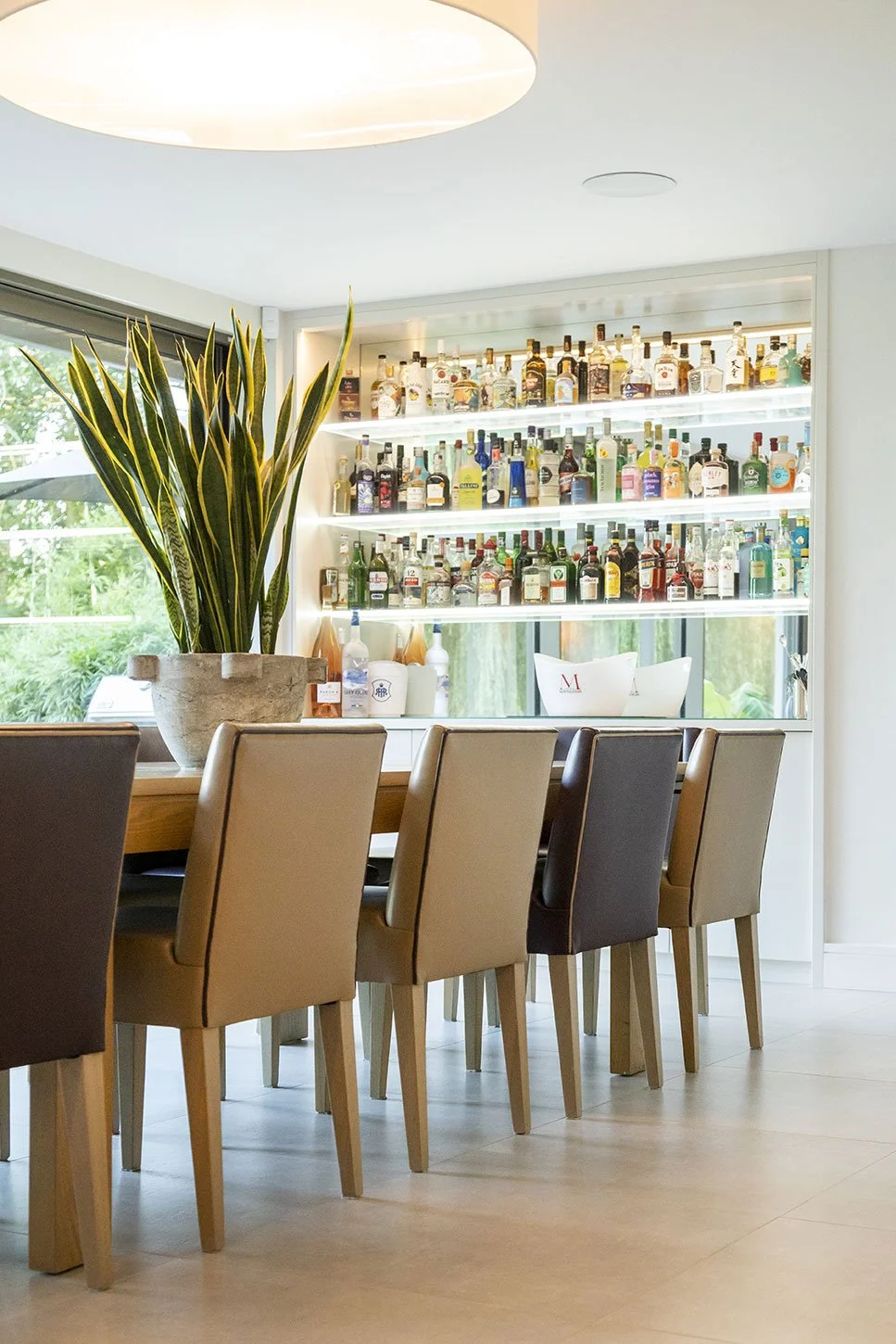 A bar area with a large potted green and yellow plant on a wooden table, beige and black chairs, and a glass-fronted liquor display with bottles on backlit shelves, next to a window with greenery outside.