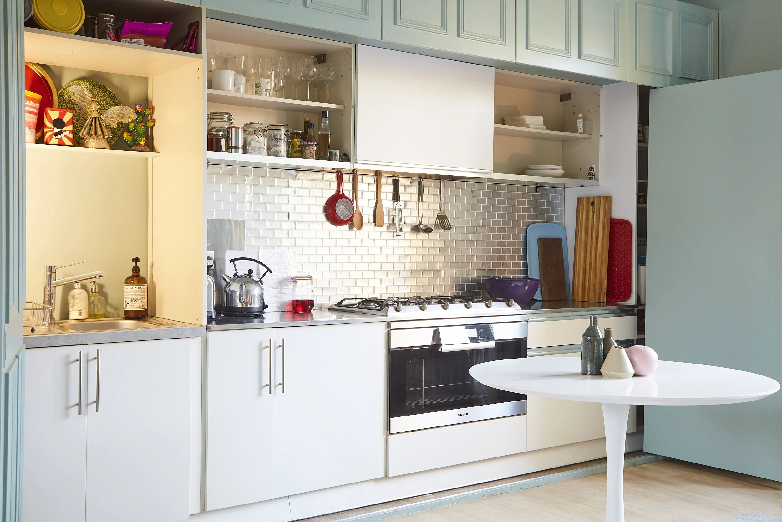 Modern kitchen with white cabinets, stainless steel appliances, open shelves with glassware and dishes, a tiled backsplash, a white round table with decorative vases, and kitchen utensils hanging above the stove.