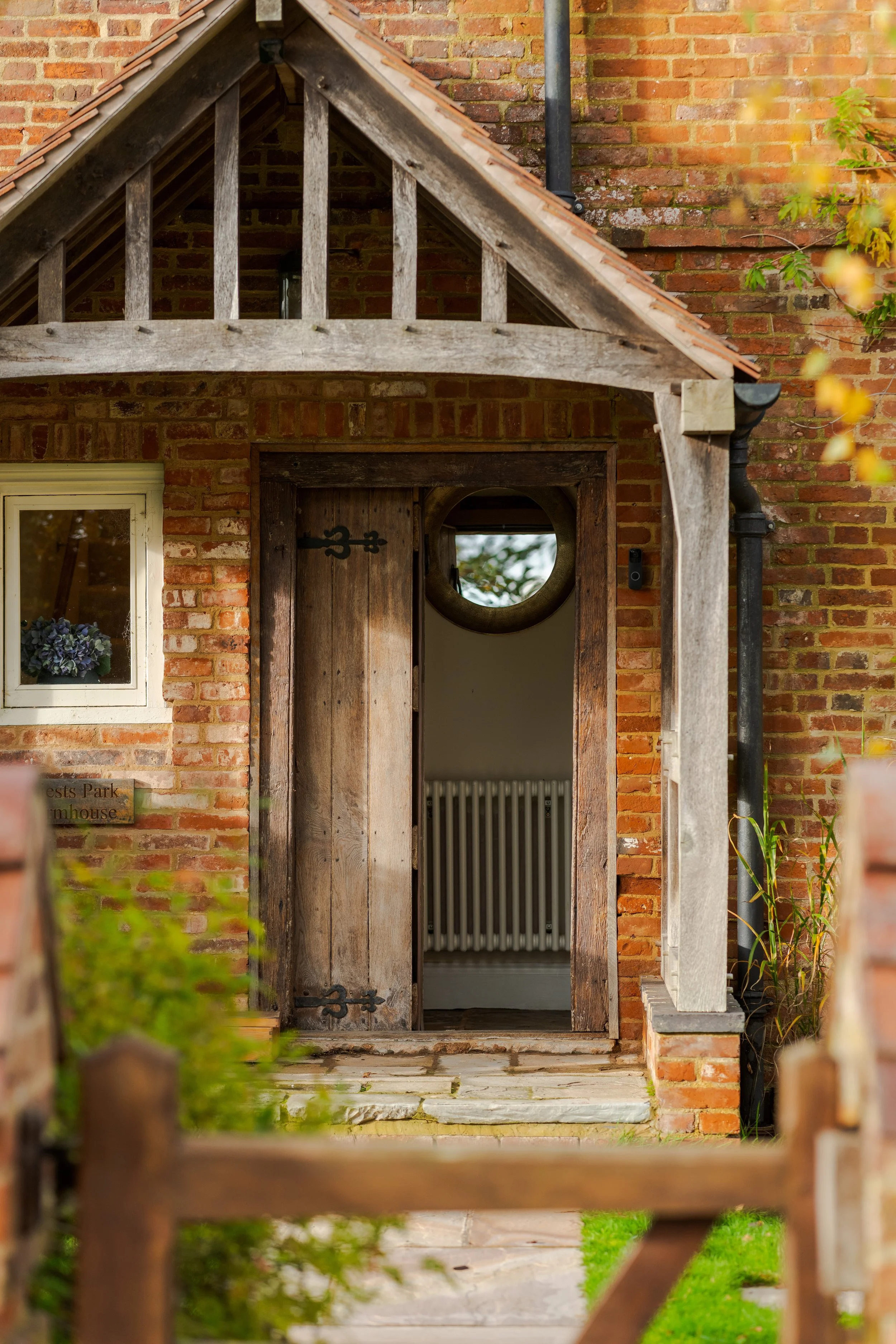 View of the entrance to a rustic brick and wood cottage with a wooden door featuring black iron hinges, a circular window, and a sign on the left that reads 'Ests Park Homhouse.' The scene includes a small stone pathway, a wooden fence in the foregro