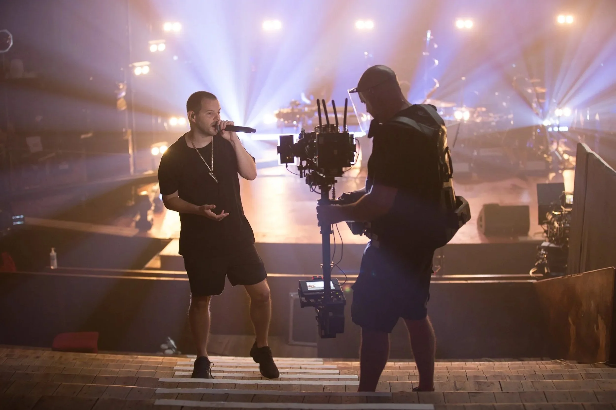 A person holding a microphone is being filmed by a cameraman on a theater stage with bright lights and a foggy atmosphere.