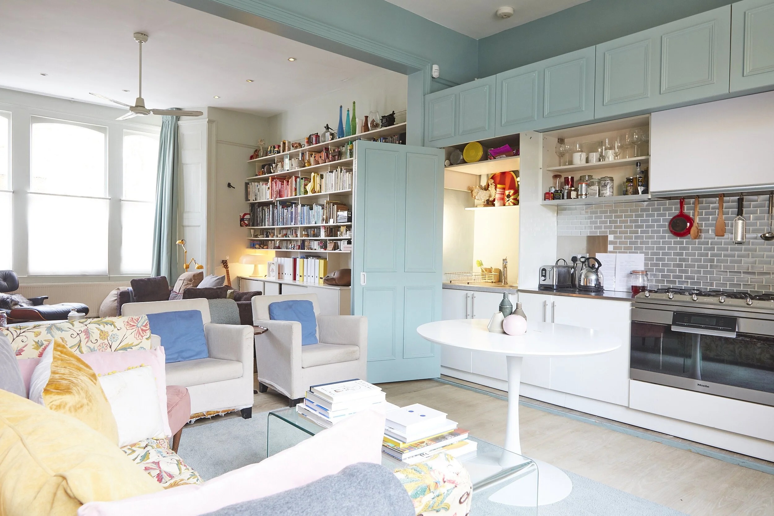 Open concept living room and kitchen with large windows, white and pastel-colored furniture, a bookshelf, and various kitchen appliances and utensils.