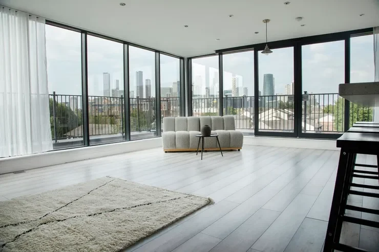 Modern living room with large floor-to-ceiling windows, a white sofa, a small side table, a beige rug, and a city skyline view.