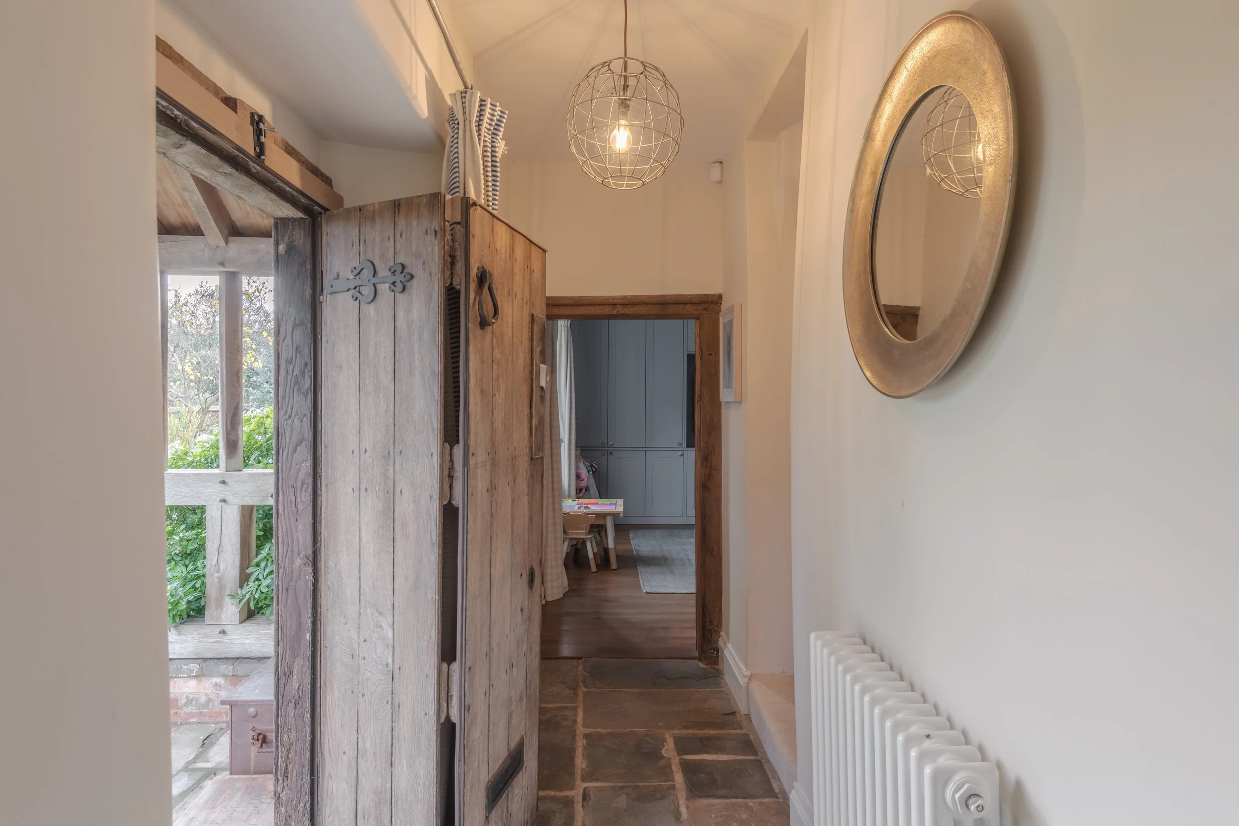 Inside view of a hallway with a rustic wooden door about to open, revealing a room with light blue cabinets, a small wooden table with chairs, a rug, and a window with curtains. A round mirror with a gold frame is mounted on the wall next to a white 