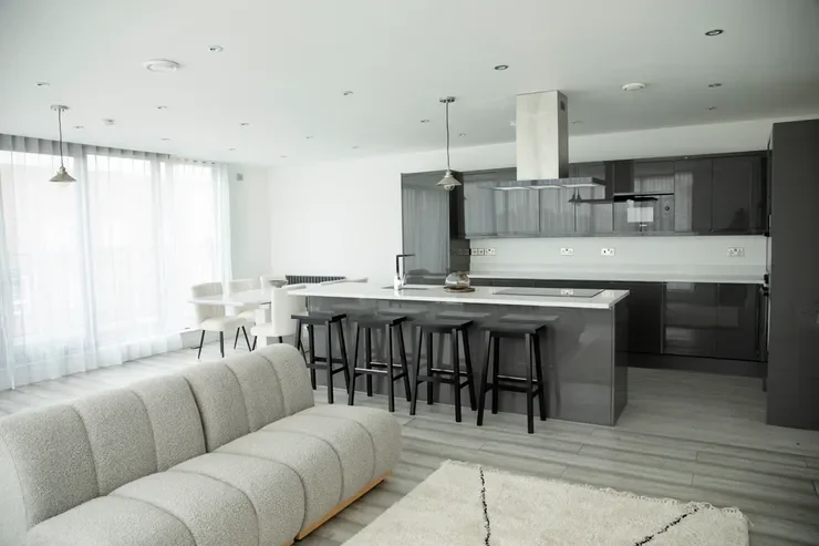 Modern open-concept kitchen and living room with gray cabinetry, a white island with four black stools, a gray sofa, and sheer white curtains.