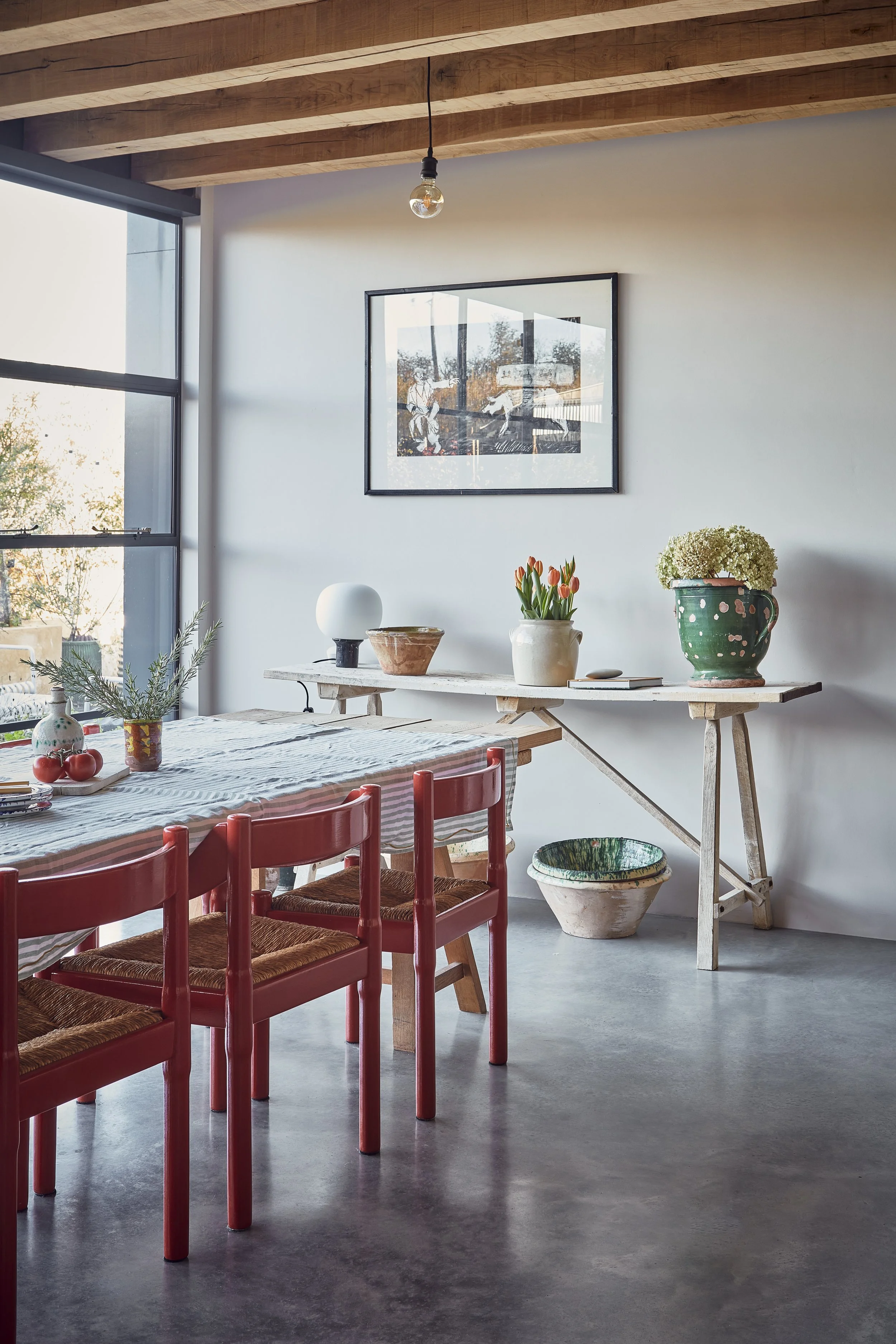Interior of a modern dining area with a wooden ceiling, large window, framed artwork on the wall, and a table with red chairs and various decorative items including plants and pottery.